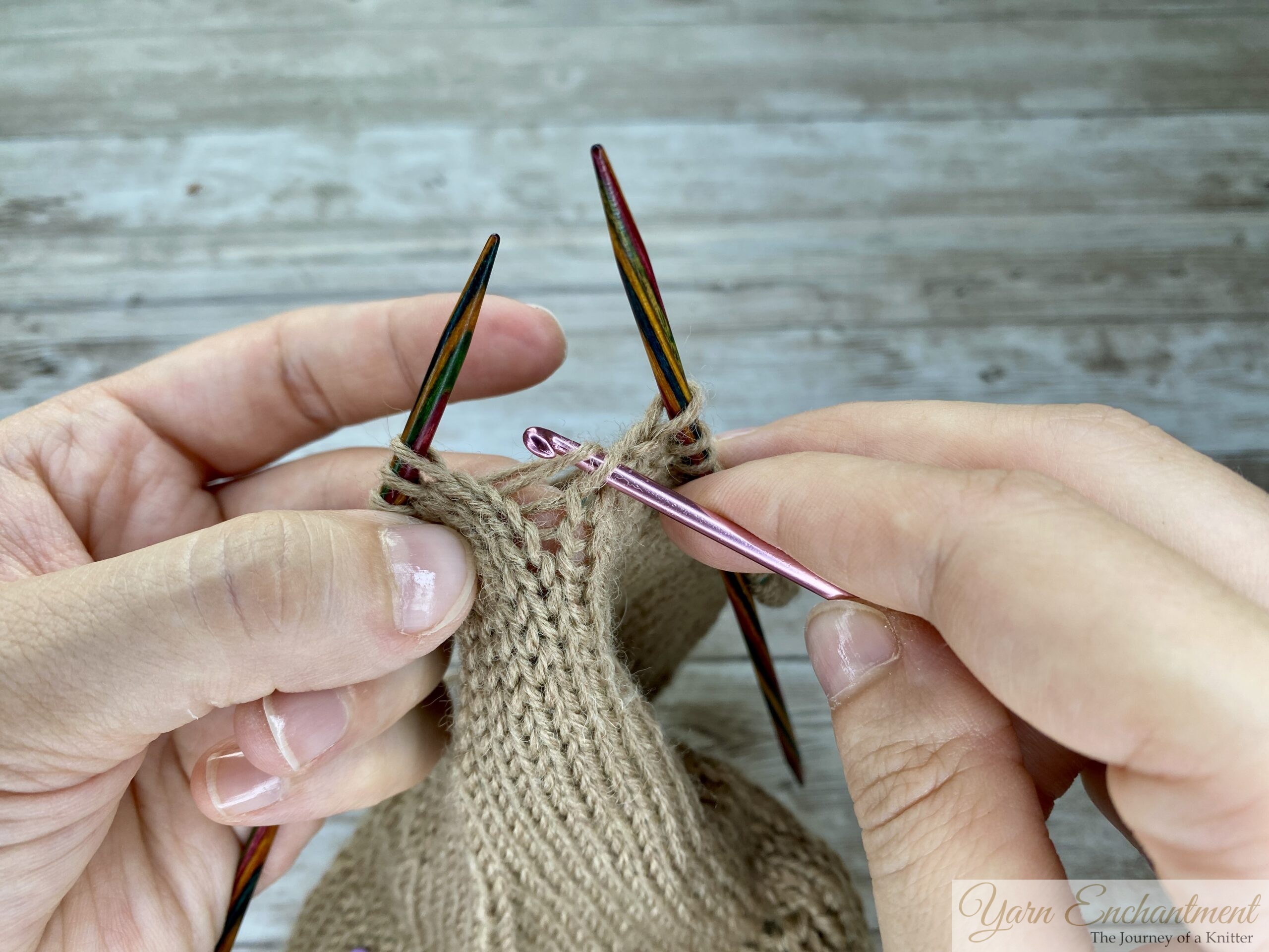 Hands using a crochet hook to pull the first horizontal ladder strand through a dropped stitch in beige stockinette fabric, demonstrating the process of making a new knit stitch.