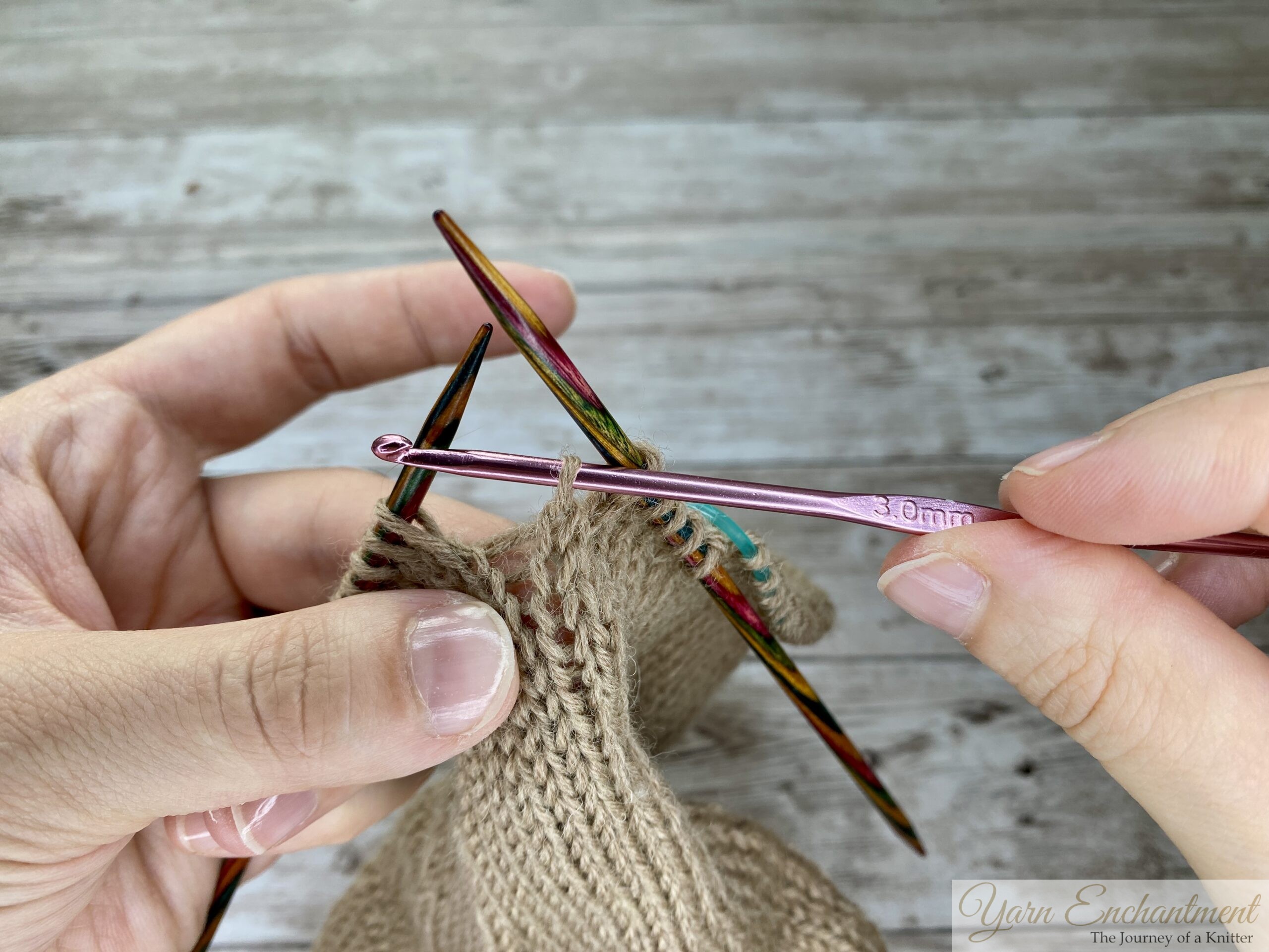 Crochet hook pulling a horizontal ladder strand through a dropped stitch in beige stockinette knitting, showing the creation of a new knit stitch with colorful double-pointed needles holding the work