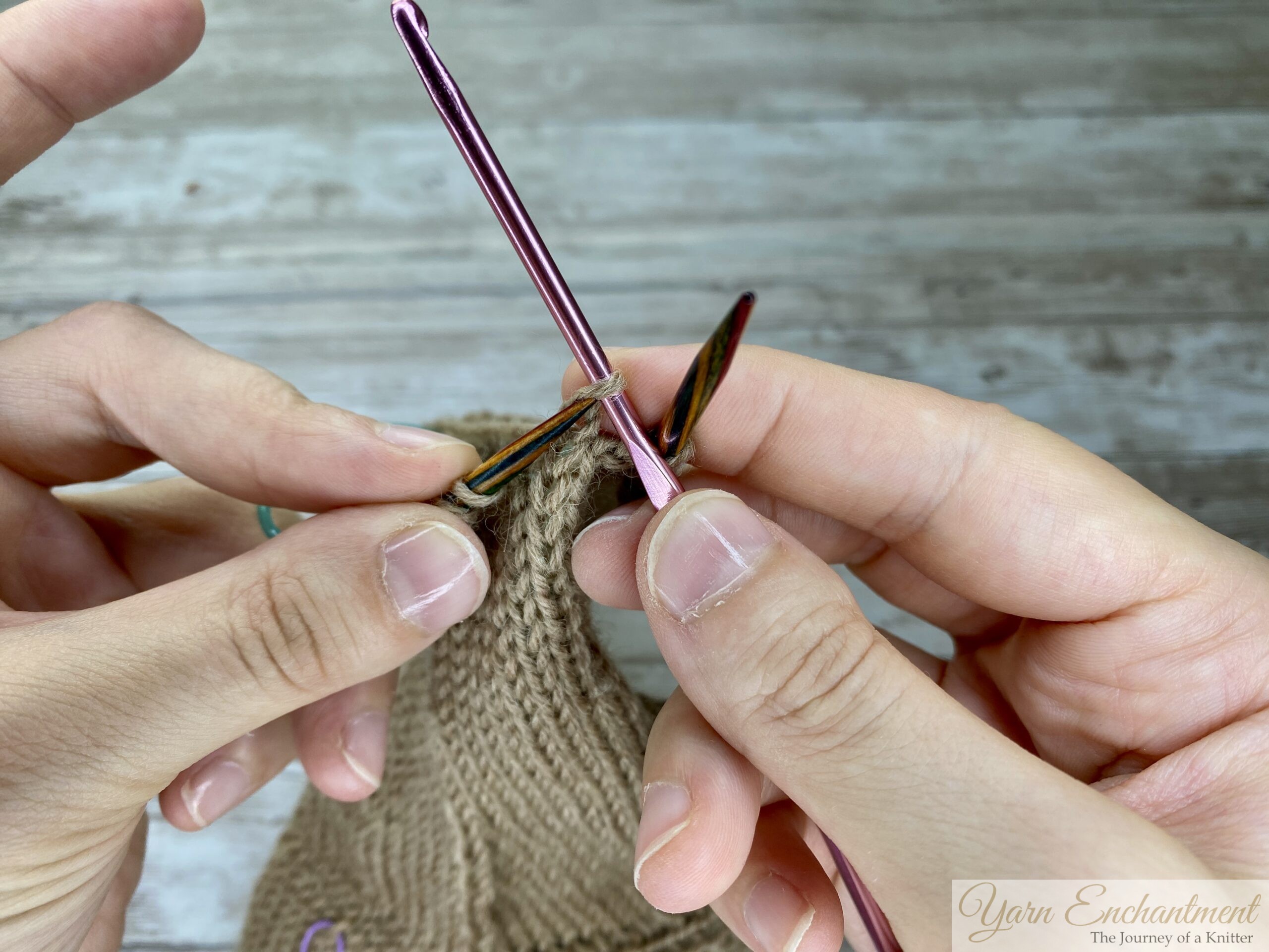 Hands using a pink crochet hook to lift the last repaired stitch in beige stockinette knitting, preparing to transfer it back onto the left knitting needle. Colorful double-pointed needles hold the remaining stitches.