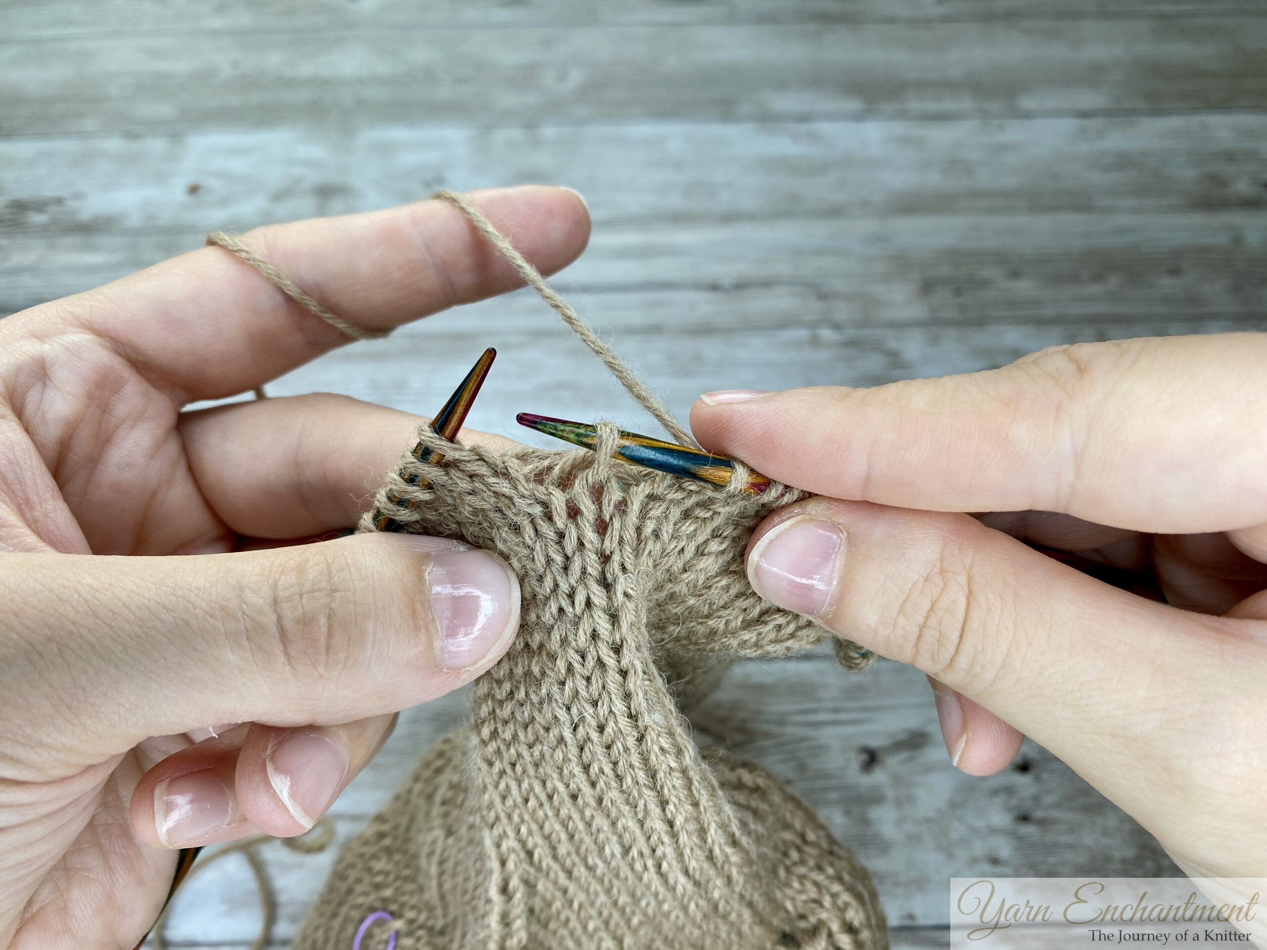Hands holding colorful knitting needles, identifying a dropped stitch in beige stockinette fabric with visible horizontal ladders above the stitch.