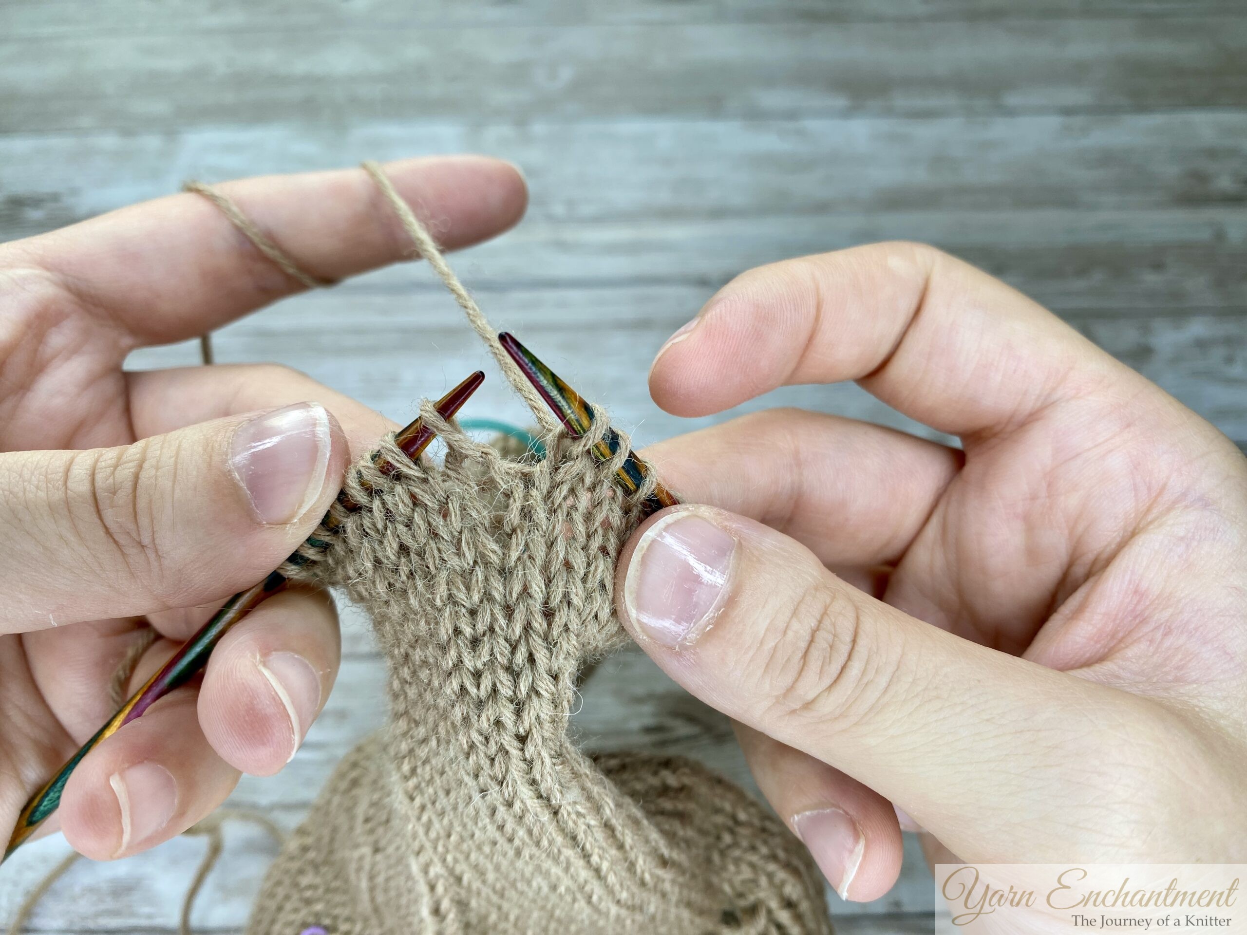 Hands returning the repaired stitch onto the left knitting needle, with all stitches aligned and ready to continue knitting.