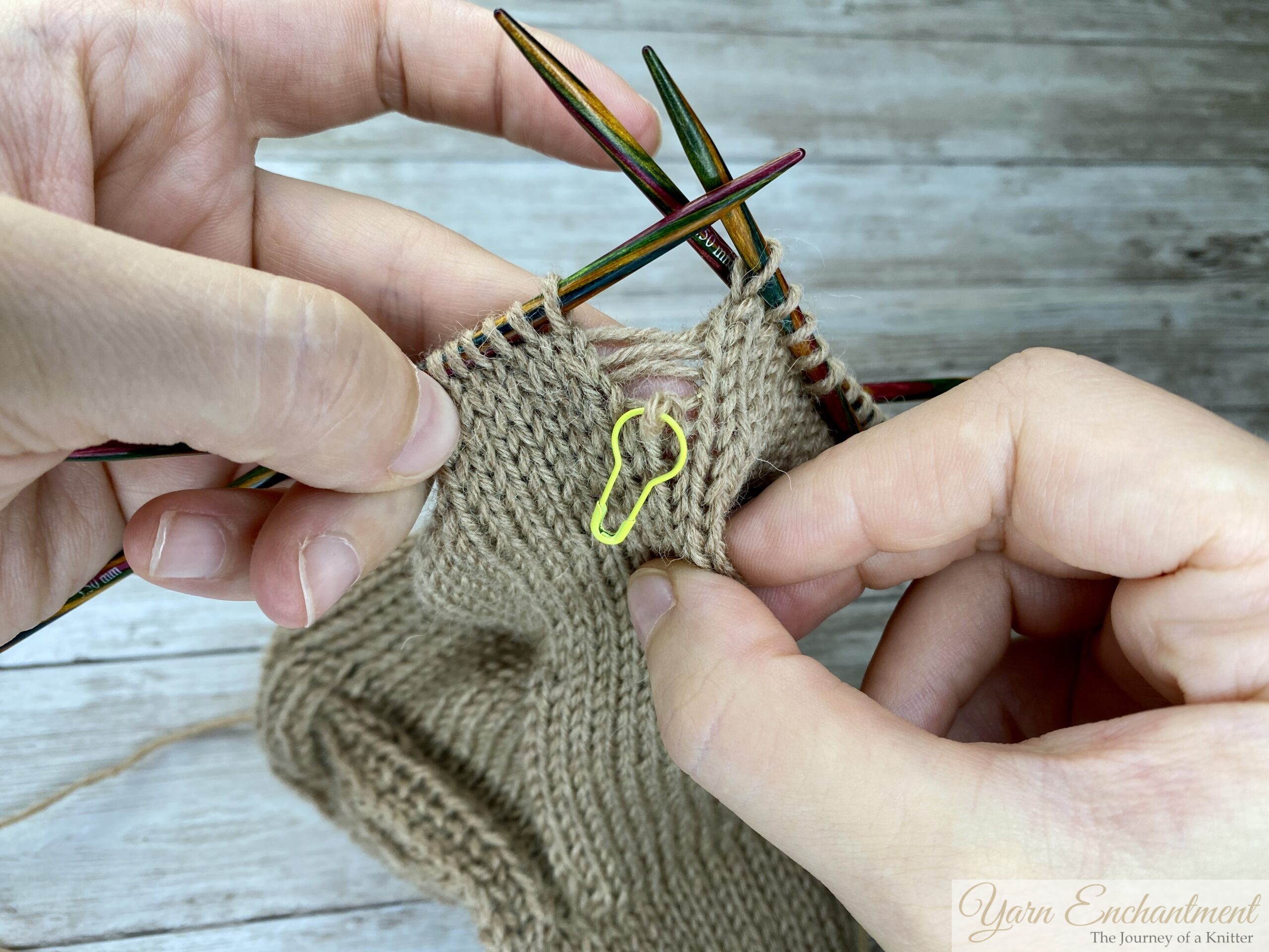 Hands holding colorful double-pointed knitting needles, demonstrating how to fix a dropped stitch in beige stockinette fabric. A yellow stitch marker highlights the dropped stitch, with several horizontal yarn strands (ladders) visible above it.