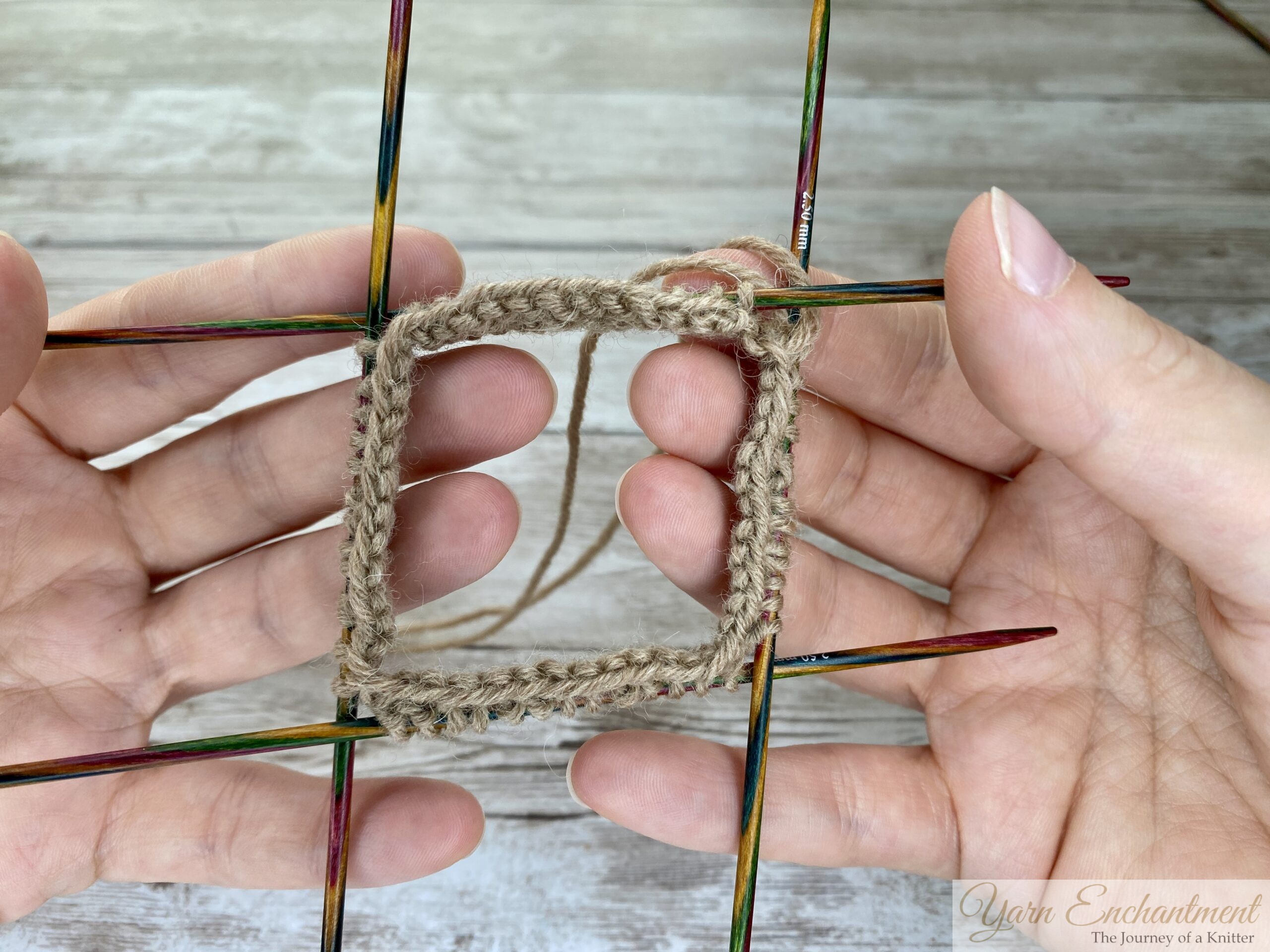 A close-up photo shows two hands holding four double-pointed knitting needles arranged in a square. Each needle holds a side of a square frame made from tan yarn, with stitches evenly distributed. The background is a light wooden surface, and the knitting appears to be at an early stage, ready for knitting in the round. No other objects are visible in the image.