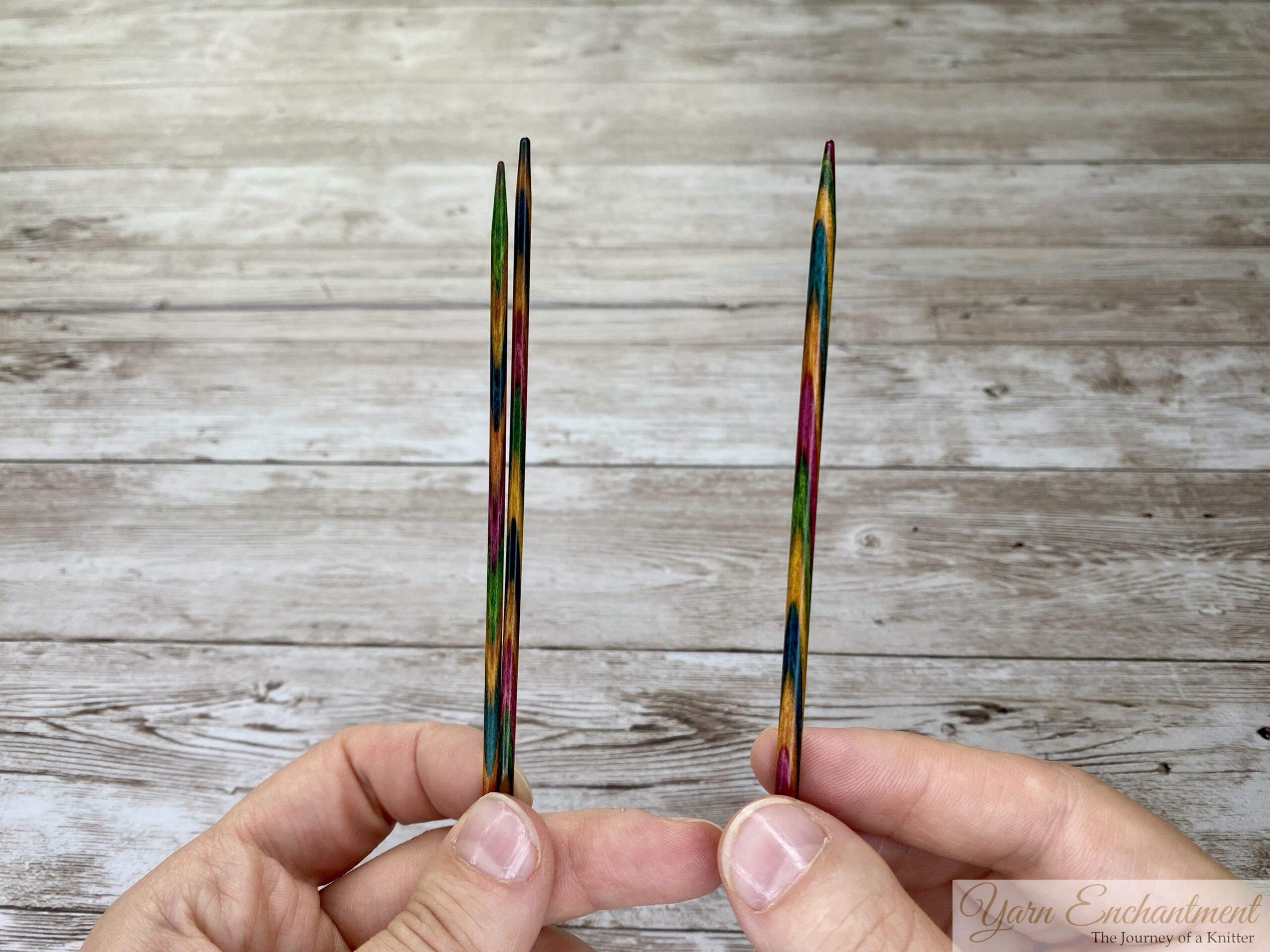 Two hands each hold a single colorful wooden knitting needle upright against a light wooden background. The needles are pointed upward and feature a striped pattern in various colors. No yarn or other objects are visible in the image.