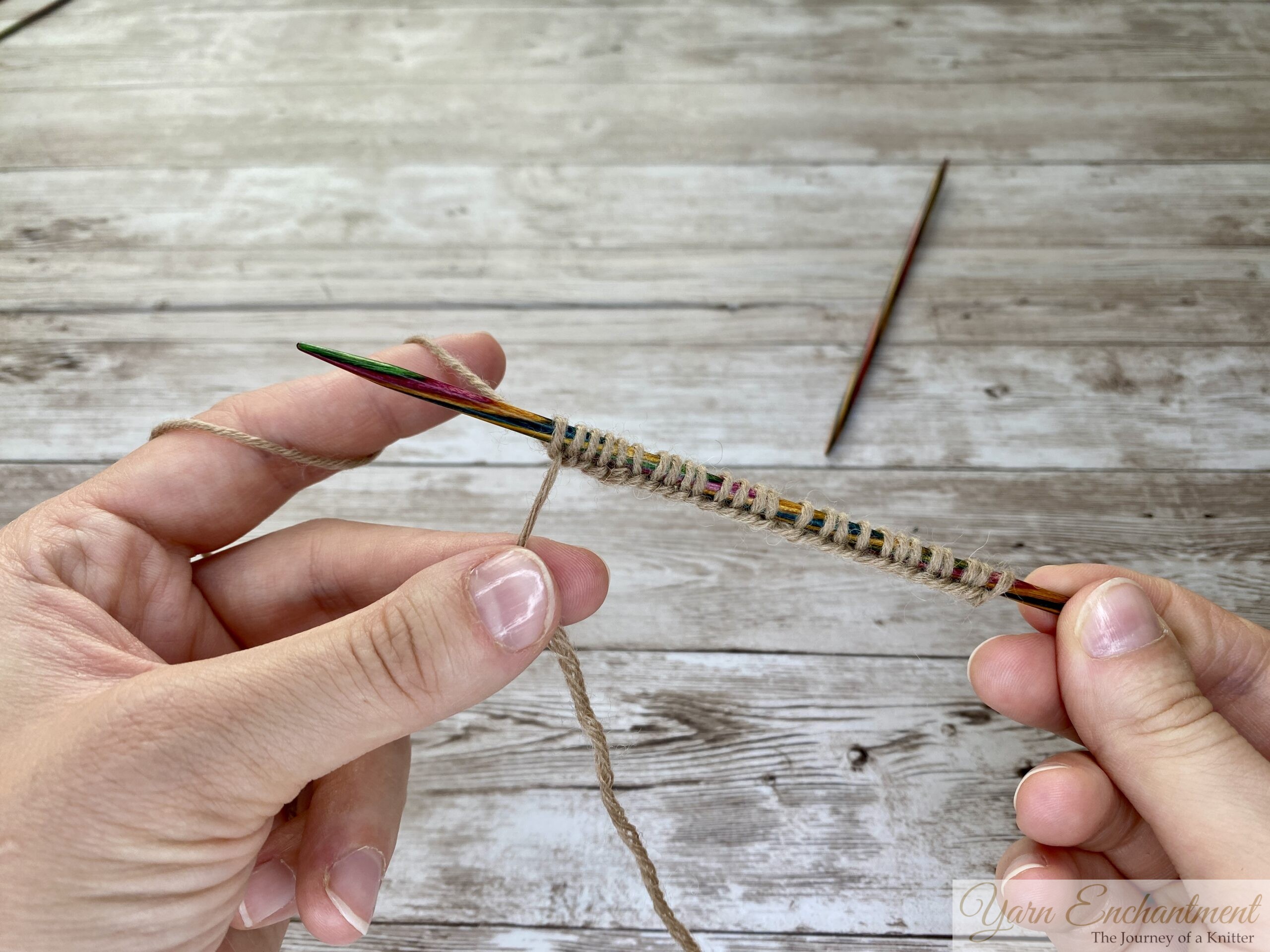 A close-up photo of two hands holding a colorful wooden knitting needle with multiple stitches of tan yarn cast onto it. The yarn is wrapped around the left hand’s fingers, and the right hand holds the needle horizontally. Another needle lies in the background on a light wooden surface.