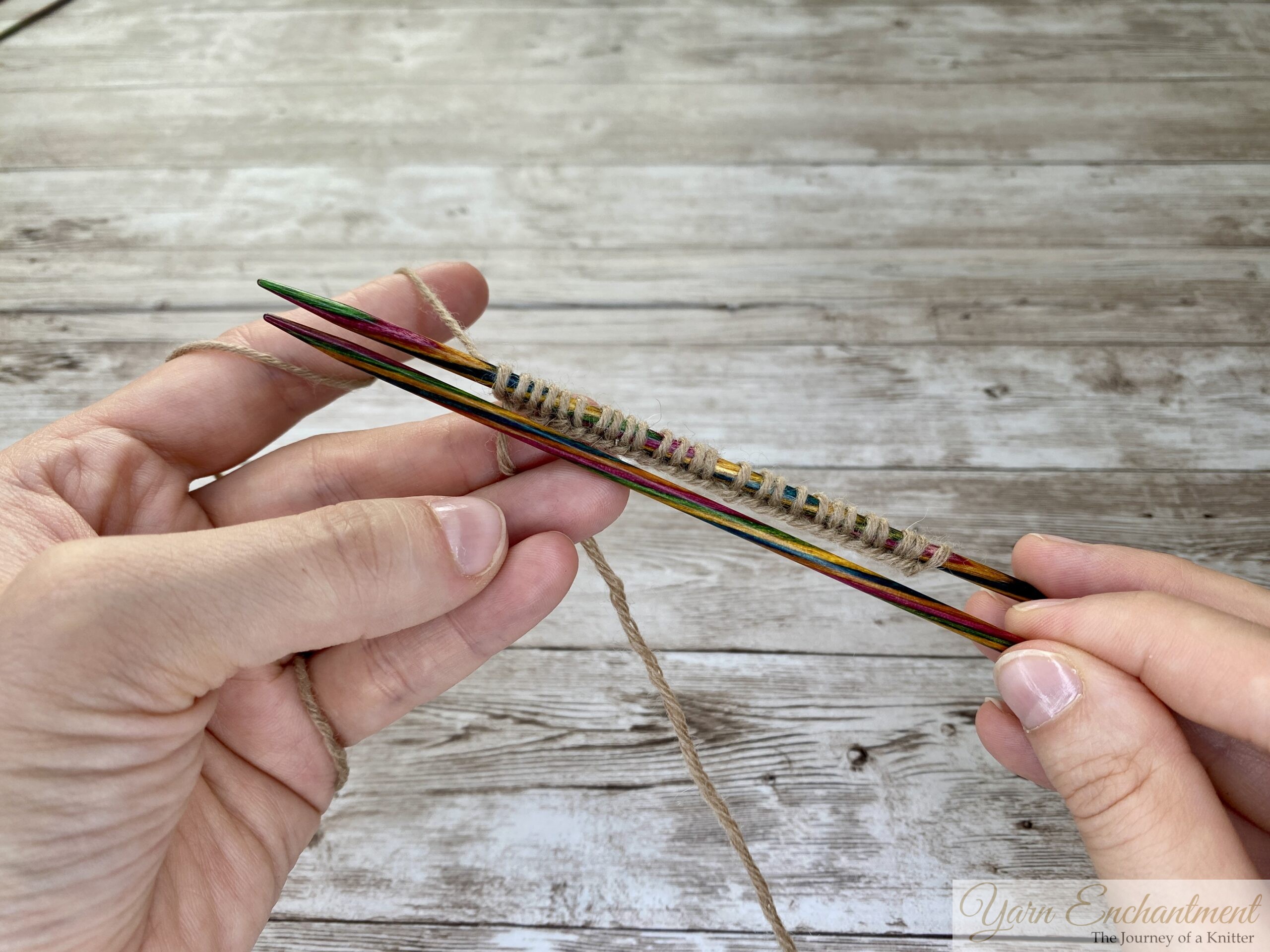 A close-up photo shows two hands holding four colorful wooden knitting needles parallel to each other. Several stitches of tan yarn are cast onto all four needles at once, with the yarn wrapped around the left hand’s fingers. The background is a light wooden surface.
