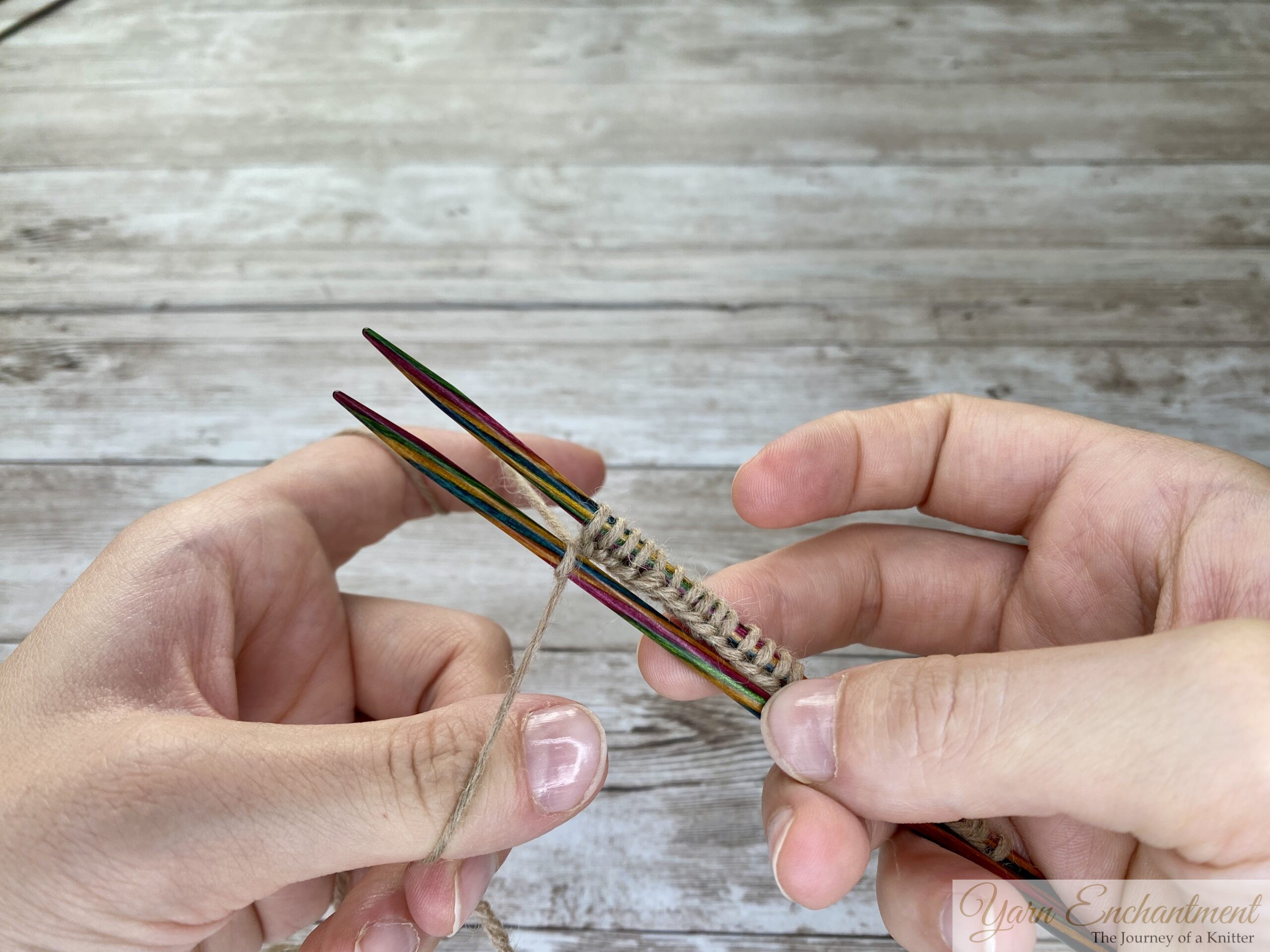 A close-up photo shows two hands holding four colorful wooden knitting needles parallel to each other. Several stitches of tan yarn are cast onto all four needles at once, with the yarn wrapped around the left hand’s fingers. The background is a light wooden surface.