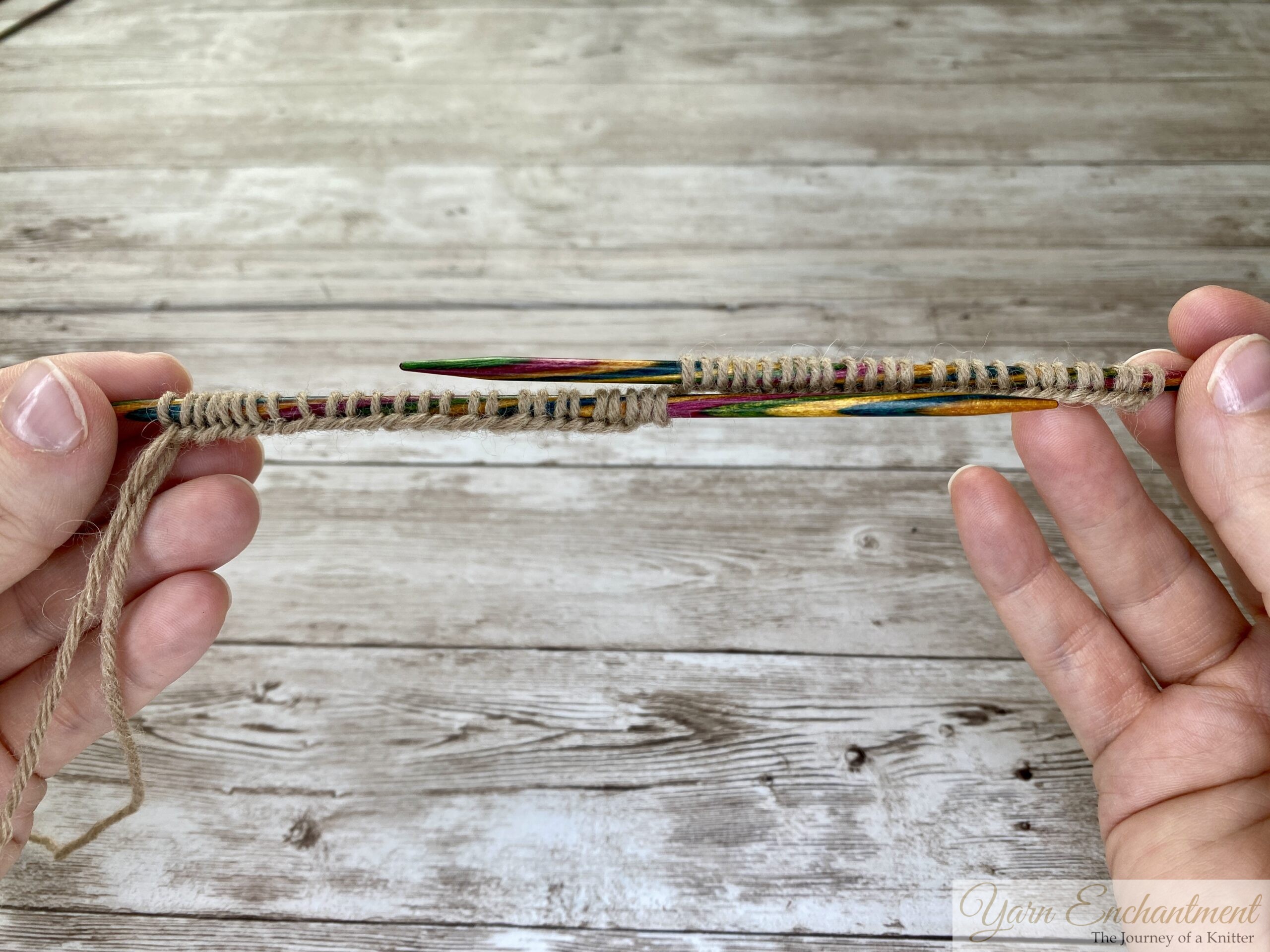A close-up photo shows two hands holding two parallel colorful wooden knitting needles. Both needles have tan yarn stitches cast onto them, with the yarn held in the left hand. The background is a light wooden surface.