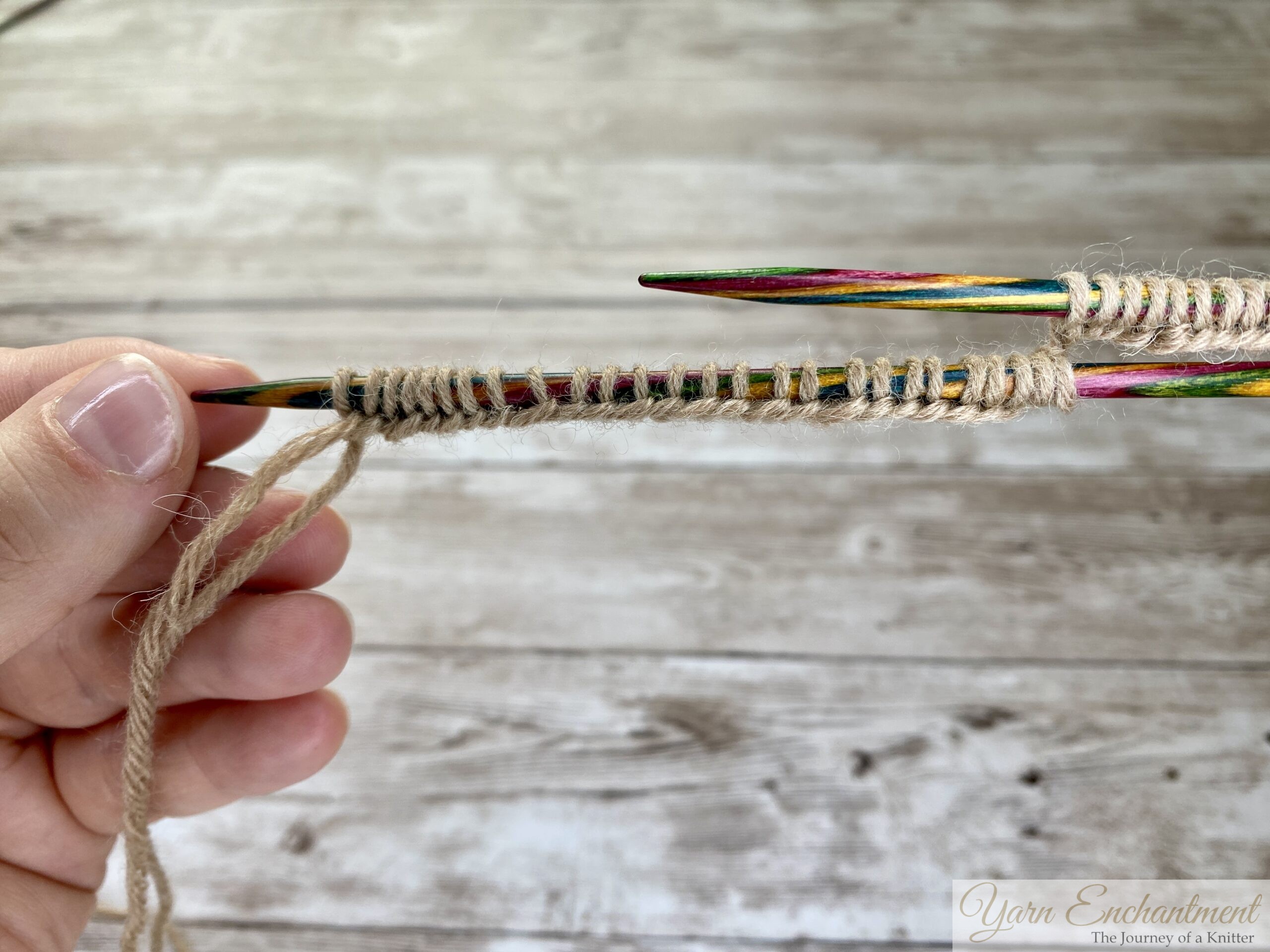 A close-up photo shows two colorful wooden knitting needles with tan yarn stitches cast onto both needles. The stitches are viewed from the right side of the knitting project, displaying neat, uniform rows. The background is a light wooden surface.