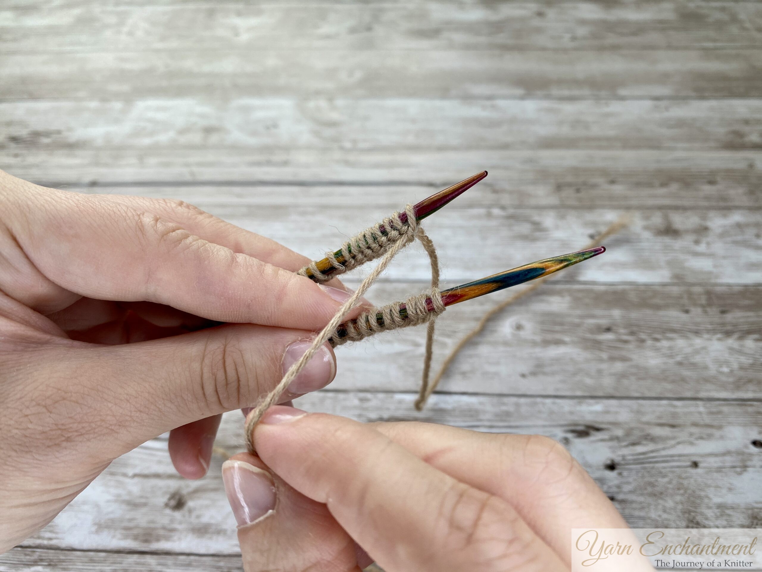 A close-up photo shows two hands holding two colorful wooden knitting needles parallel to each other. Both needles have tan yarn stitches cast onto them, and the yarn is looped around the left hand’s fingers. The background is a light wooden surface.