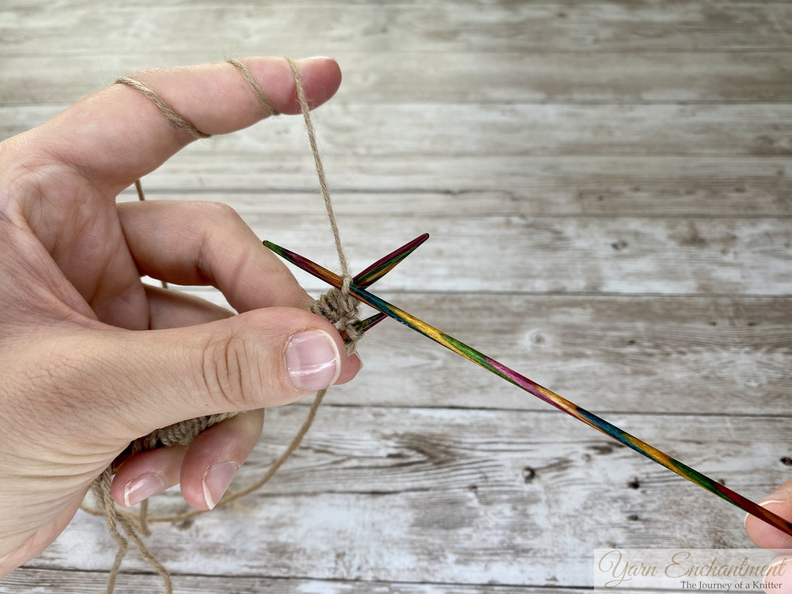 A close-up photo shows two hands holding two colorful wooden knitting needles in a V shape. Each needle has tan yarn stitches, and the yarn is looped around the left hand’s fingers. The background is a light wooden surface