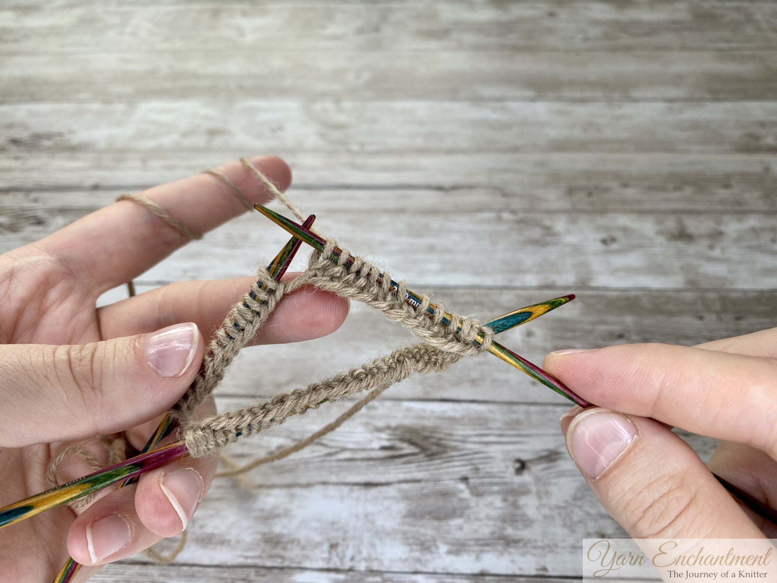 A close-up photo of two hands holding four colorful wooden double-pointed knitting needles arranged in a triangle. Tan yarn stitches are distributed evenly across the needles, and the working yarn is looped around the left hand’s fingers. The background is a light wooden surface.