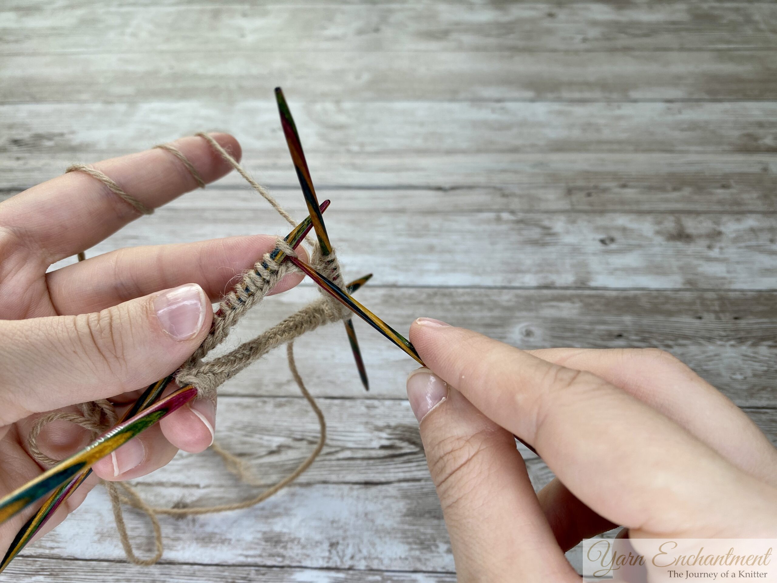 A close-up photo shows two hands holding four colorful wooden double-pointed knitting needles in a triangular arrangement. The needles have tan yarn stitches, and the working yarn is wrapped around the left hand’s fingers. The background is a light wooden surface.