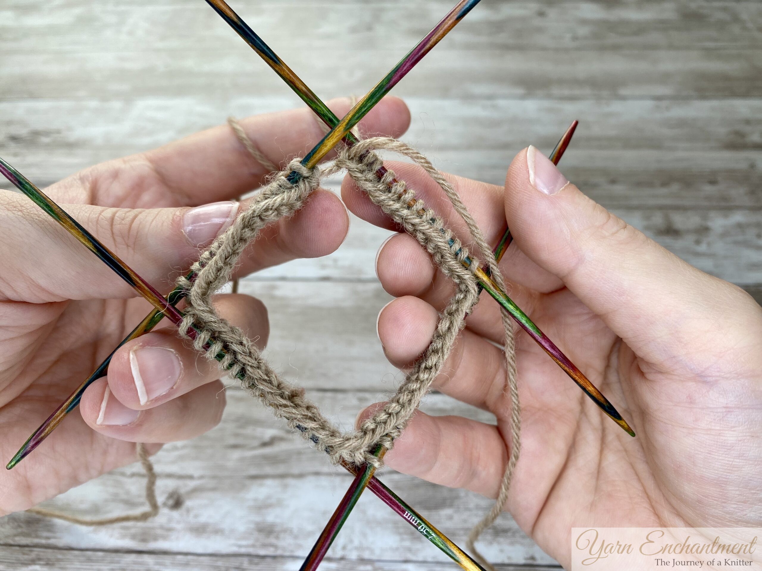 A close-up photo shows two hands holding four double-pointed knitting needles arranged in a square. Each needle holds a side of a square frame made from tan yarn, with stitches evenly distributed. The background is a light wooden surface, and the knitting appears to be at an early stage, ready for knitting in the round. No other objects are visible in the image.