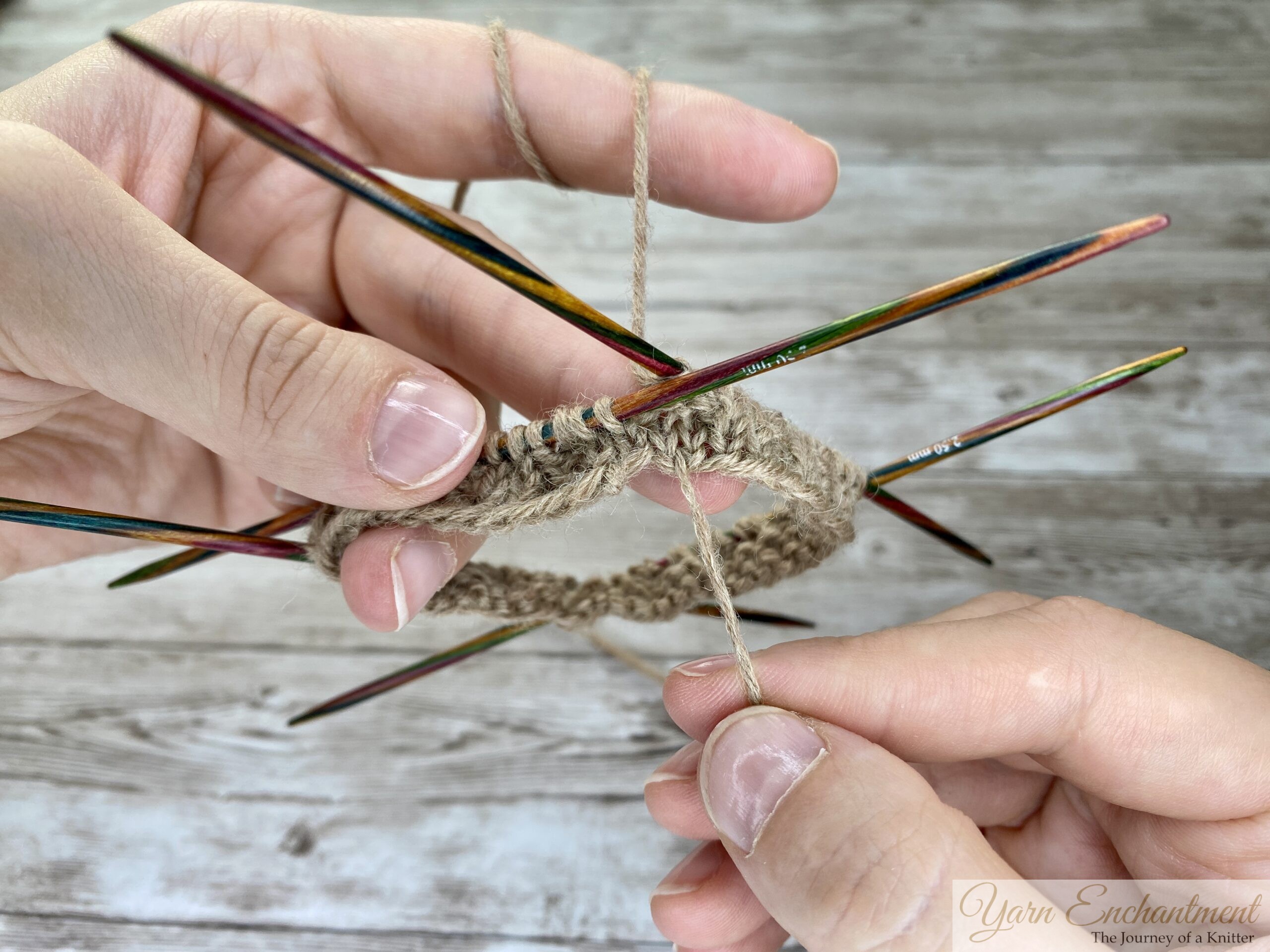 A close-up photo shows two hands holding four colorful wooden double-pointed knitting needles arranged in a square. Each needle has tan yarn stitches cast onto it, forming a circular shape. The right hand is pulling the yarn taut, preparing to knit in the round. The background is a light wooden surface.