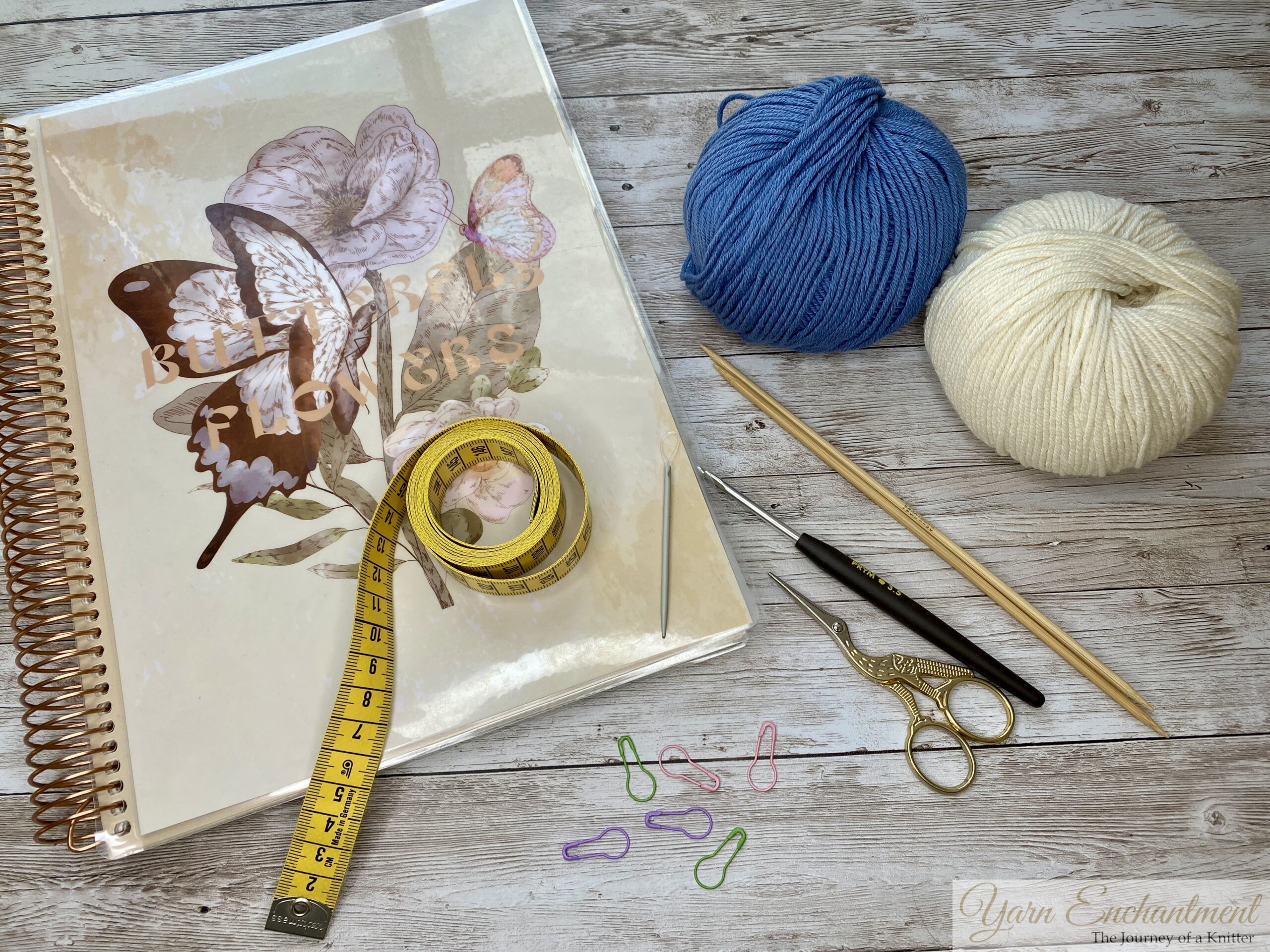 Overhead view of knitting and crochet supplies laid out on a wooden surface, including a floral notebook, a yellow tape measure, blue and white yarn balls, knitting needles, a crochet hook, gold embroidery scissors, and colorful stitch markers.