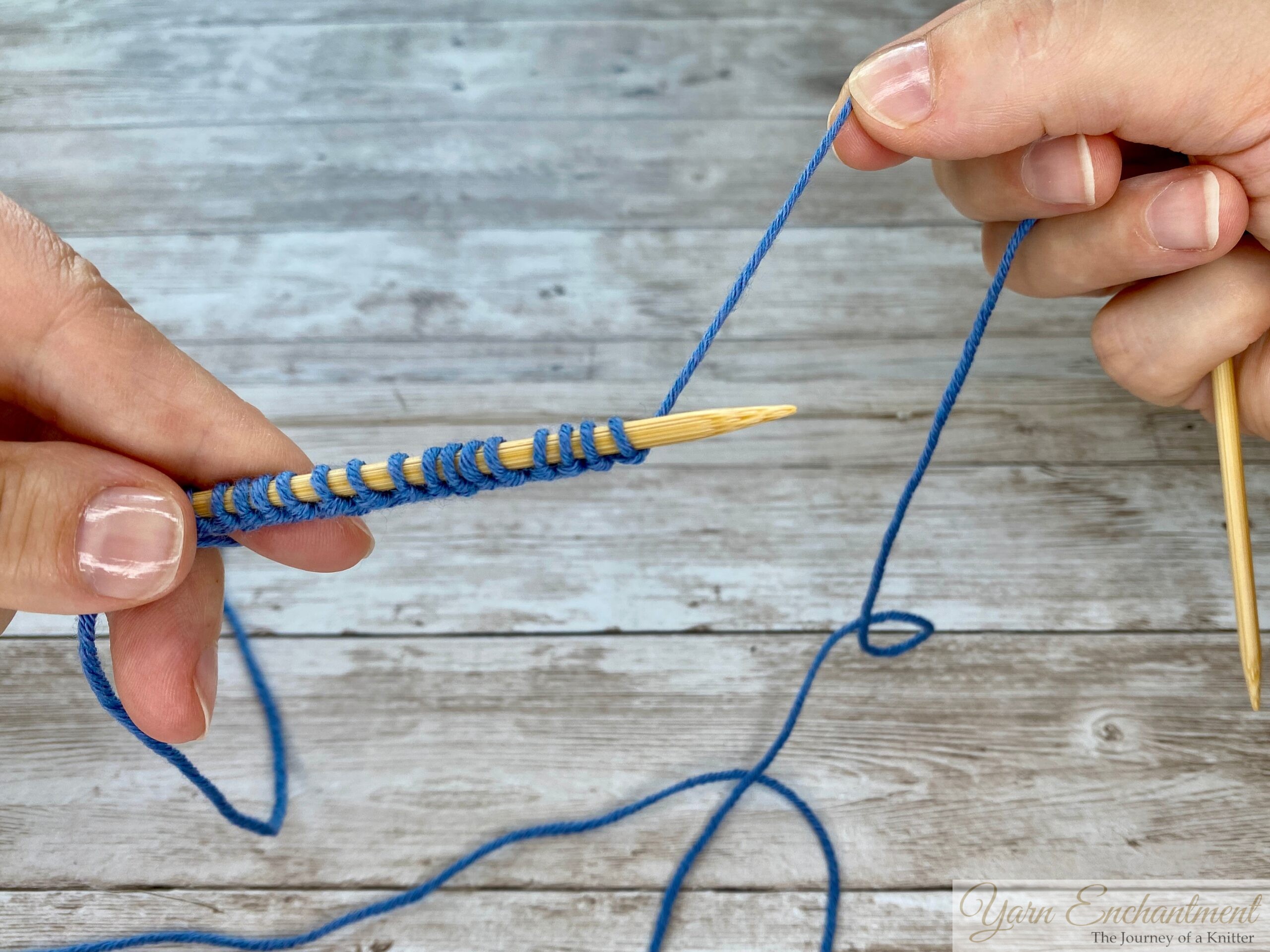 Hands displaying the completed cast-on row of blue stitches along a bamboo knitting needle, with the right needle empty and ready to continue the knitting process.