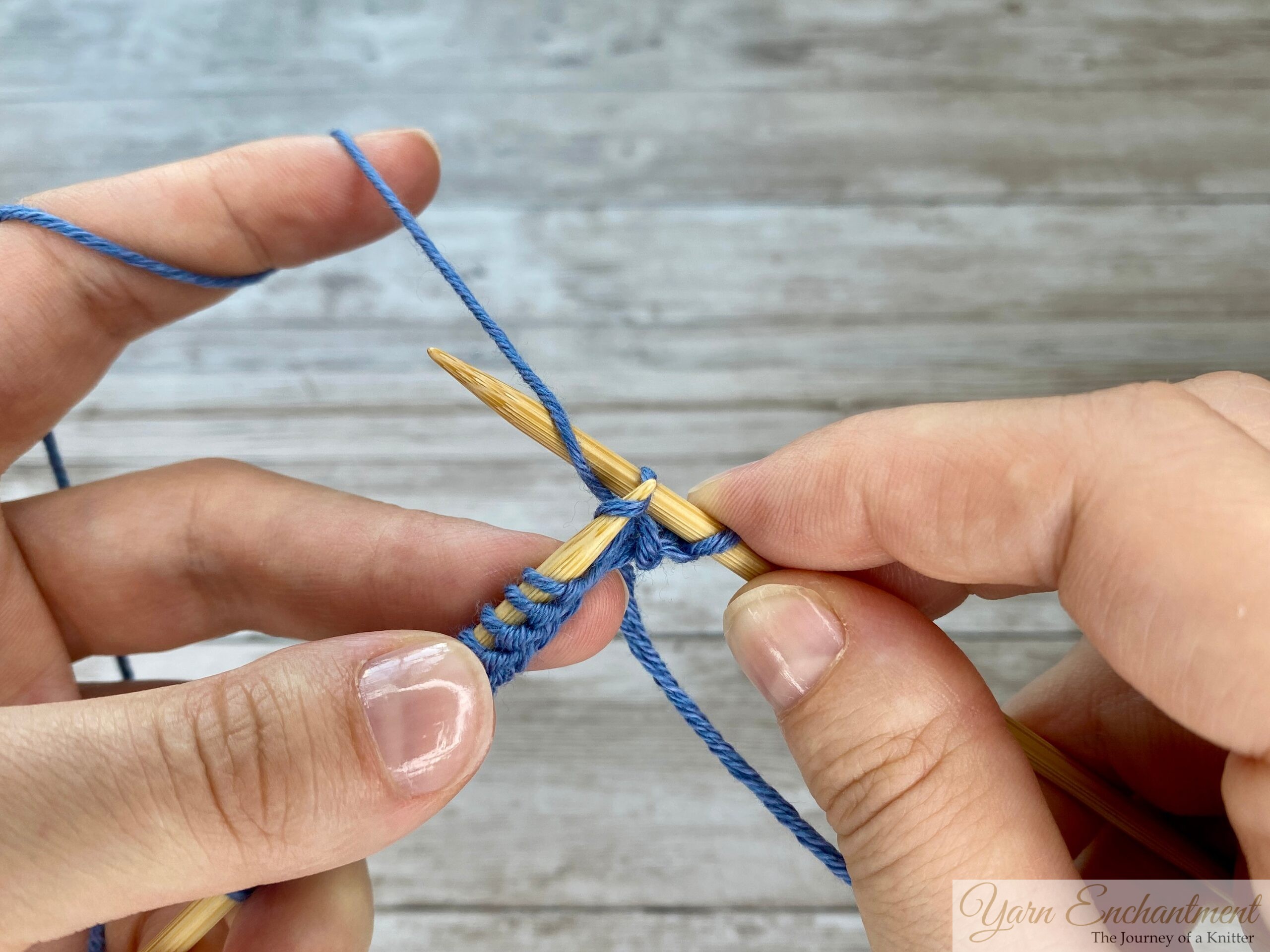 Hands displaying the next step of the knit stitch, with the working yarn wrapped around the right needle and ready to be pulled through the stitch.