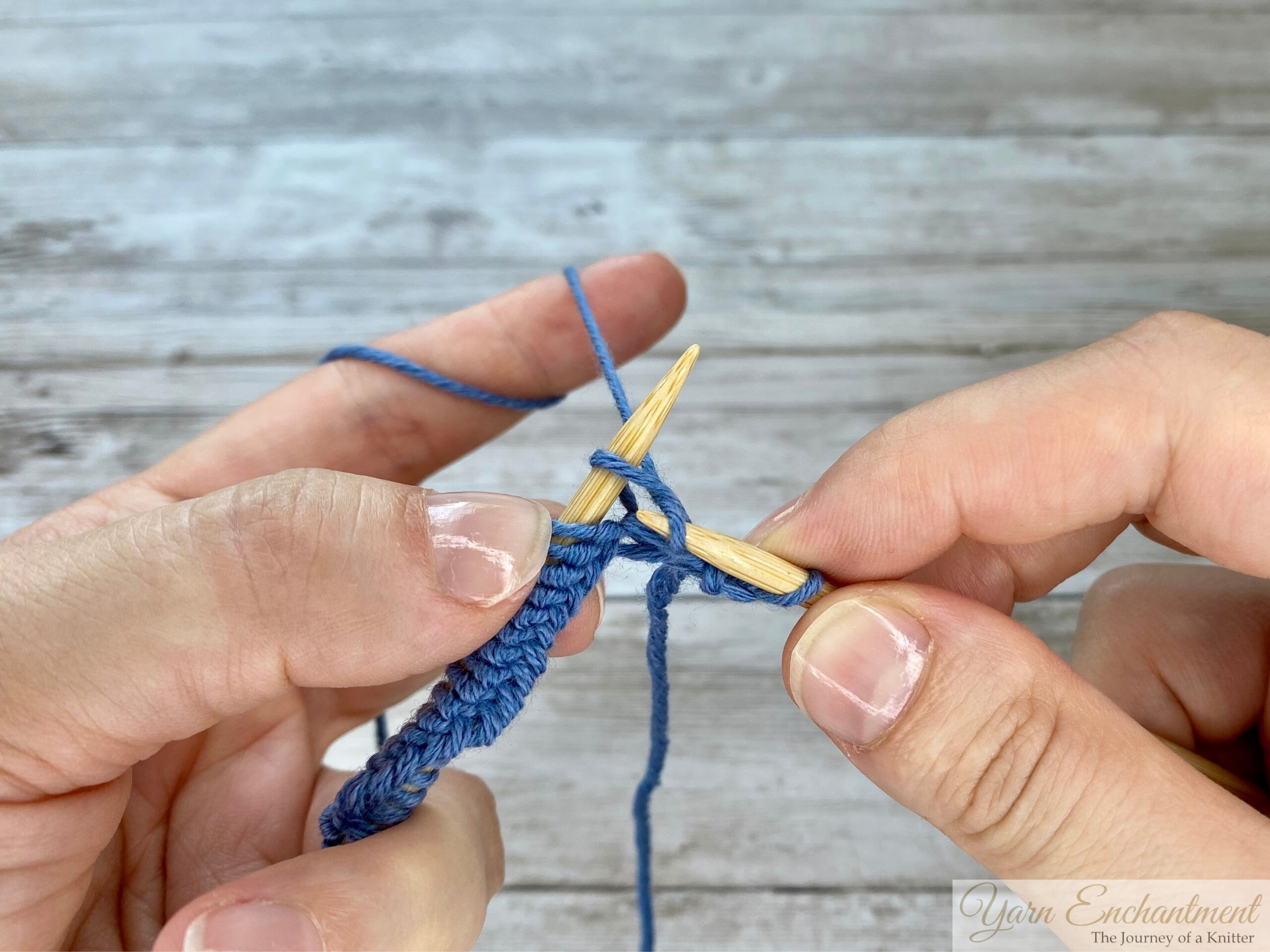 Close-up of hands with wooden knitting needles, showing the right needle inserted into the stitch as the working yarn is positioned for creating a knit stitch.