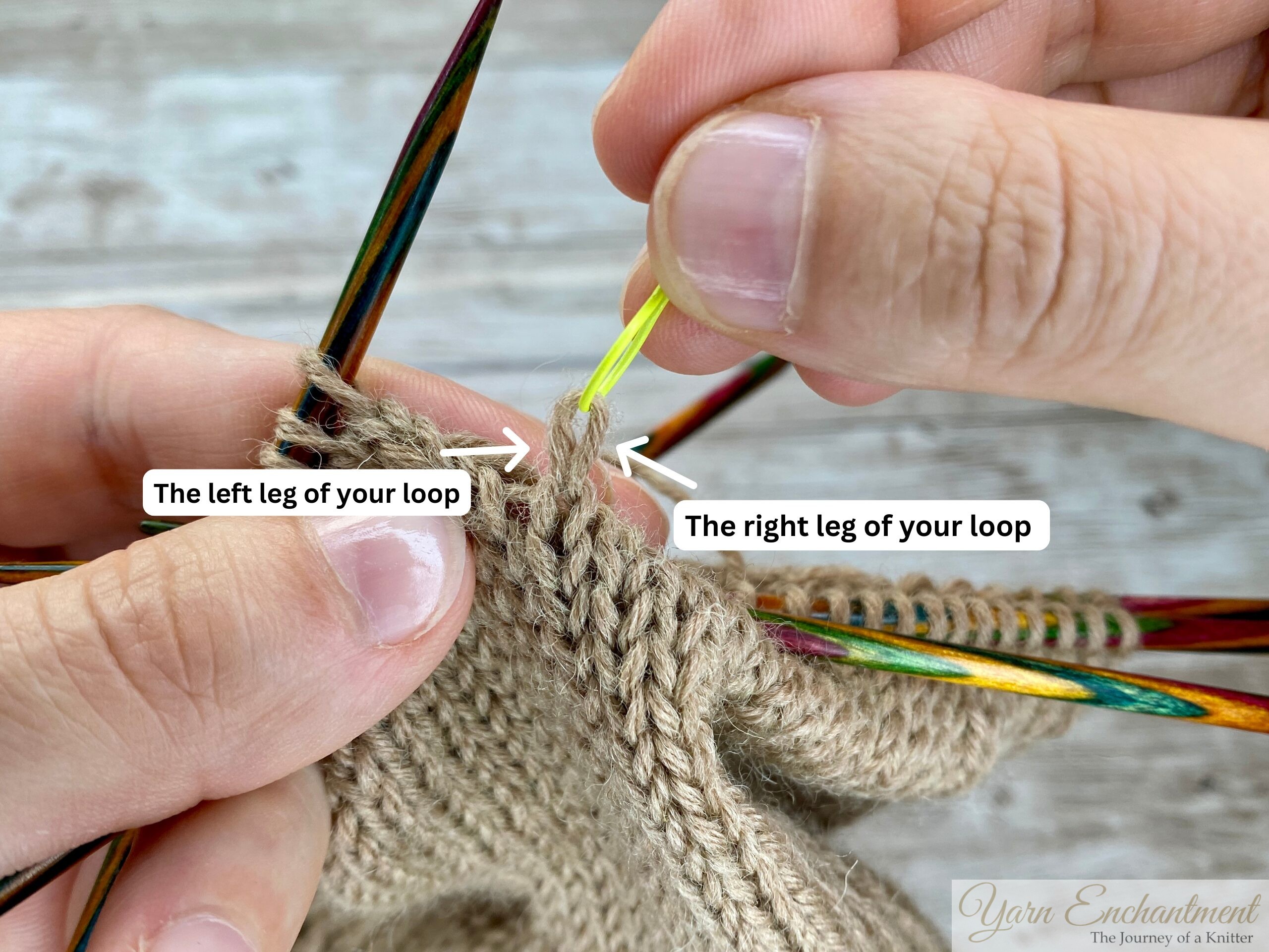 Close-up of hands using a yellow stitch marker to lift a dropped stitch in beige stockinette knitting. Colorful double-pointed needles hold the surrounding stitches, and horizontal yarn strands (ladders) are clearly visible above the dropped stitch.