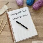 A flat lay photo of a knitting-themed workspace. On a wooden table are three skeins of yarn (one purple, one blue, one pink), a small swatch of white knitting on needles, two loose wooden knitting needles, and an open spiral notebook. The notebook’s lined page displays the handwritten title, “Knitting Skill Levels—Where Do You Fit In?” with a black and gold fountain pen resting across the page. The composition suggests a cozy, creative setting for knitting and writing.