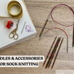 Assorted sock knitting tools arranged on a wooden table, including circular knitting needles, double-pointed needles, measuring tape, scissors, stitch markers, tapestry needle, row counter, and a notebook, with the text 'NEEDLES & ACCESSORIES FOR SOCK KNITTING.