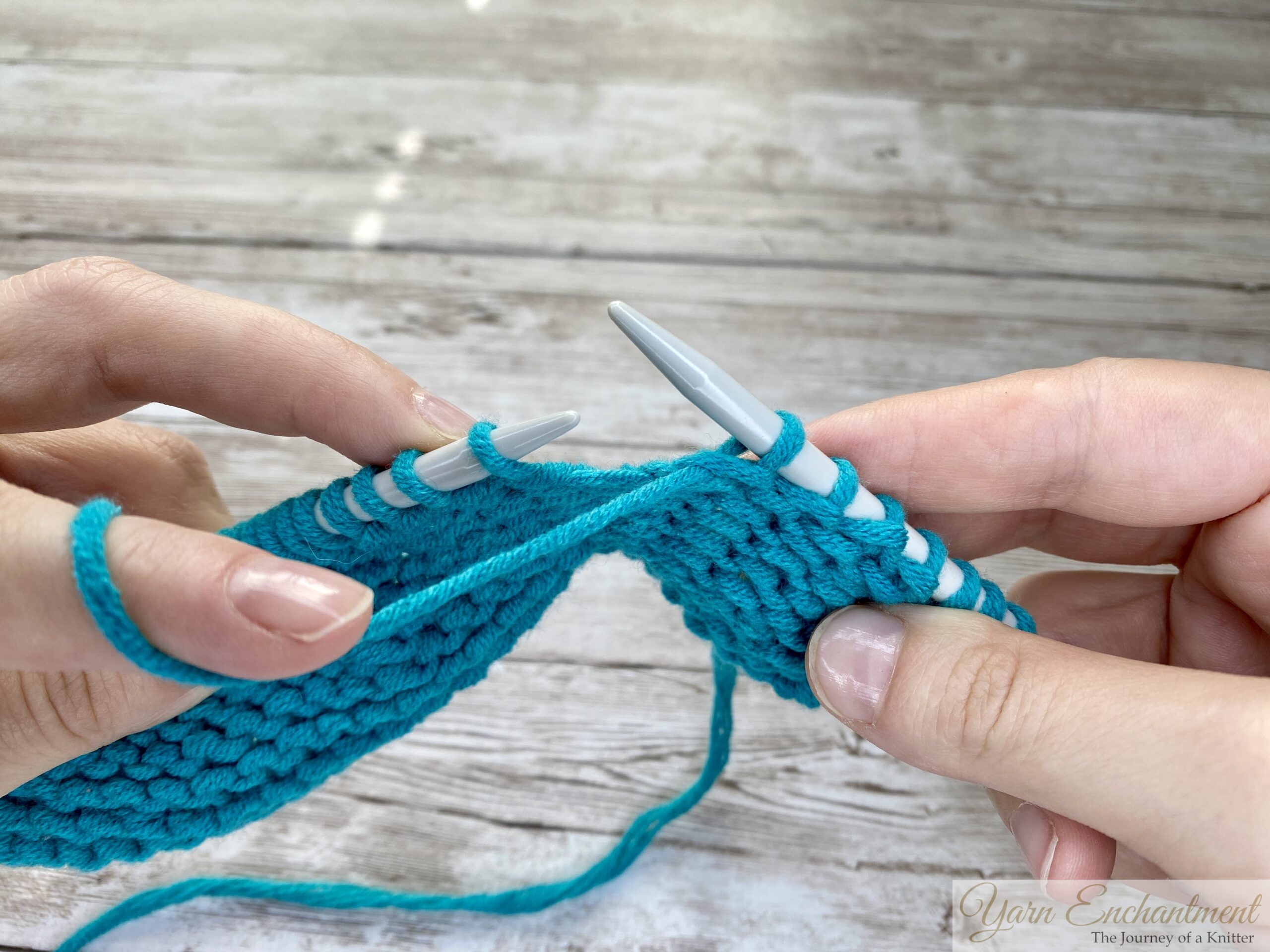 Close-up of hands demonstrating the purl stitch while knitting with turquoise yarn on white knitting needles, against a light wooden background