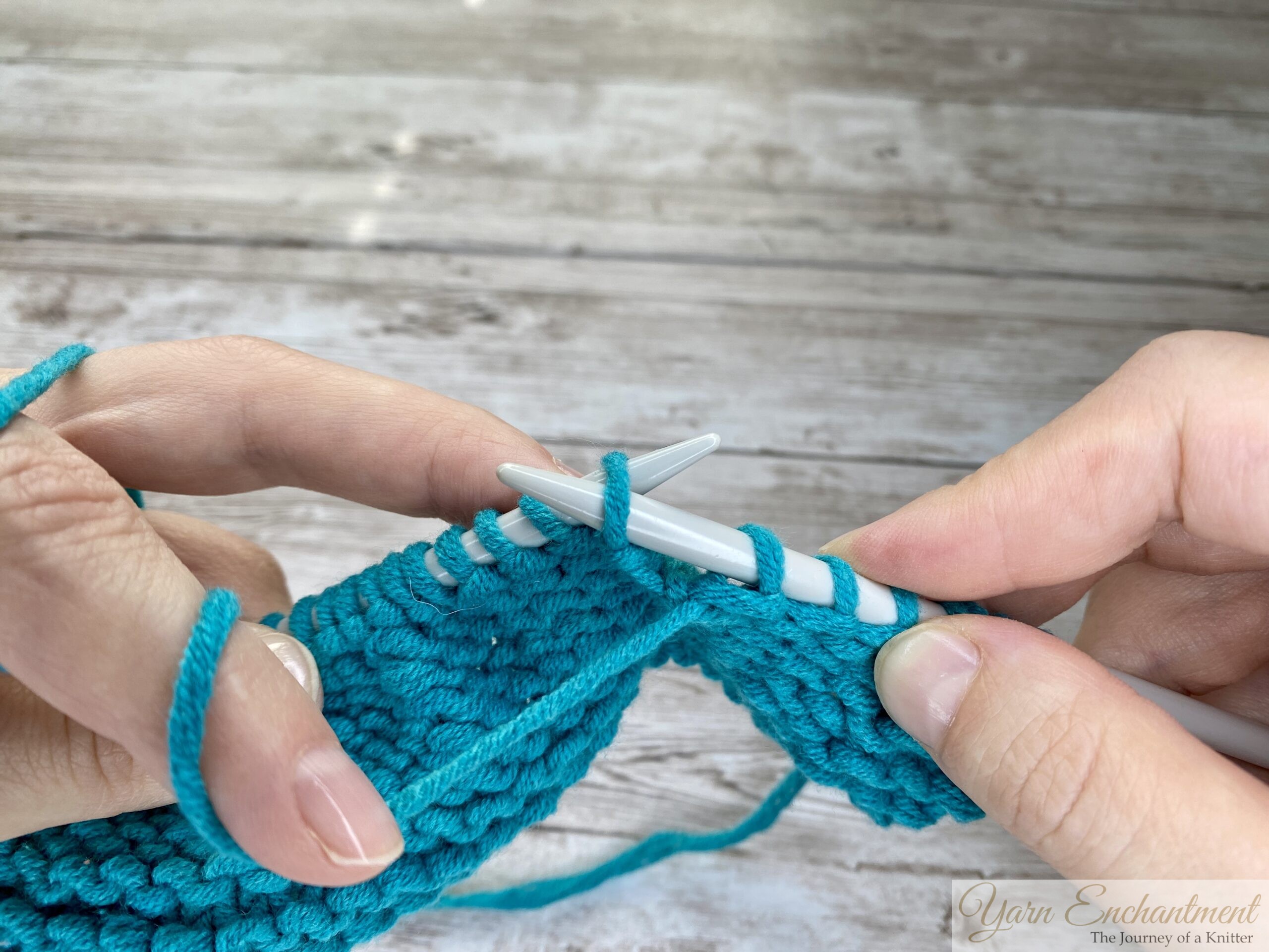 Close-up of hands performing the purl stitch with turquoise yarn and light gray knitting needles, showing the yarn in front and needles positioned to complete a stitch on a wooden surface background.