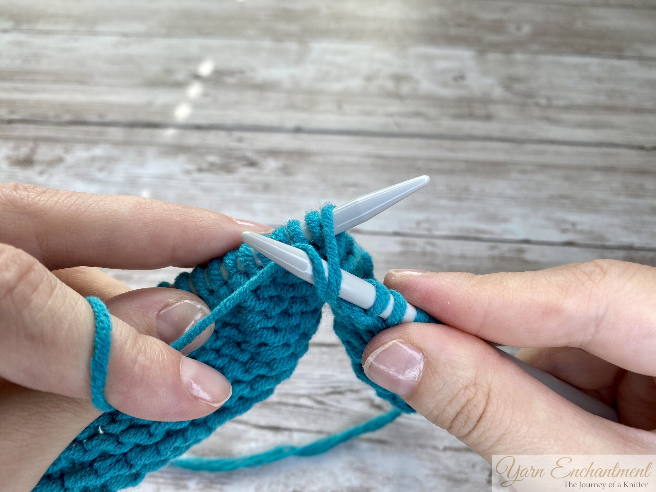 Close-up of hands pulling the right-hand knitting needle with turquoise yarn wrapped around it back through the stitch on the left-hand needle, creating a new loop to form the purl stitch.
