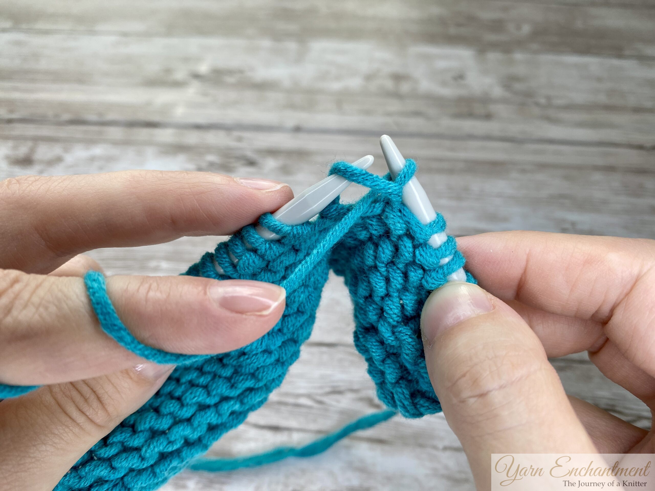 Close-up of hands carefully pulling the right-hand knitting needle back through the stitch on the left-hand needle, then sliding the old stitch off the left-hand needle after pulling the new loop through to complete the first purl stitch, all on a wooden surface.