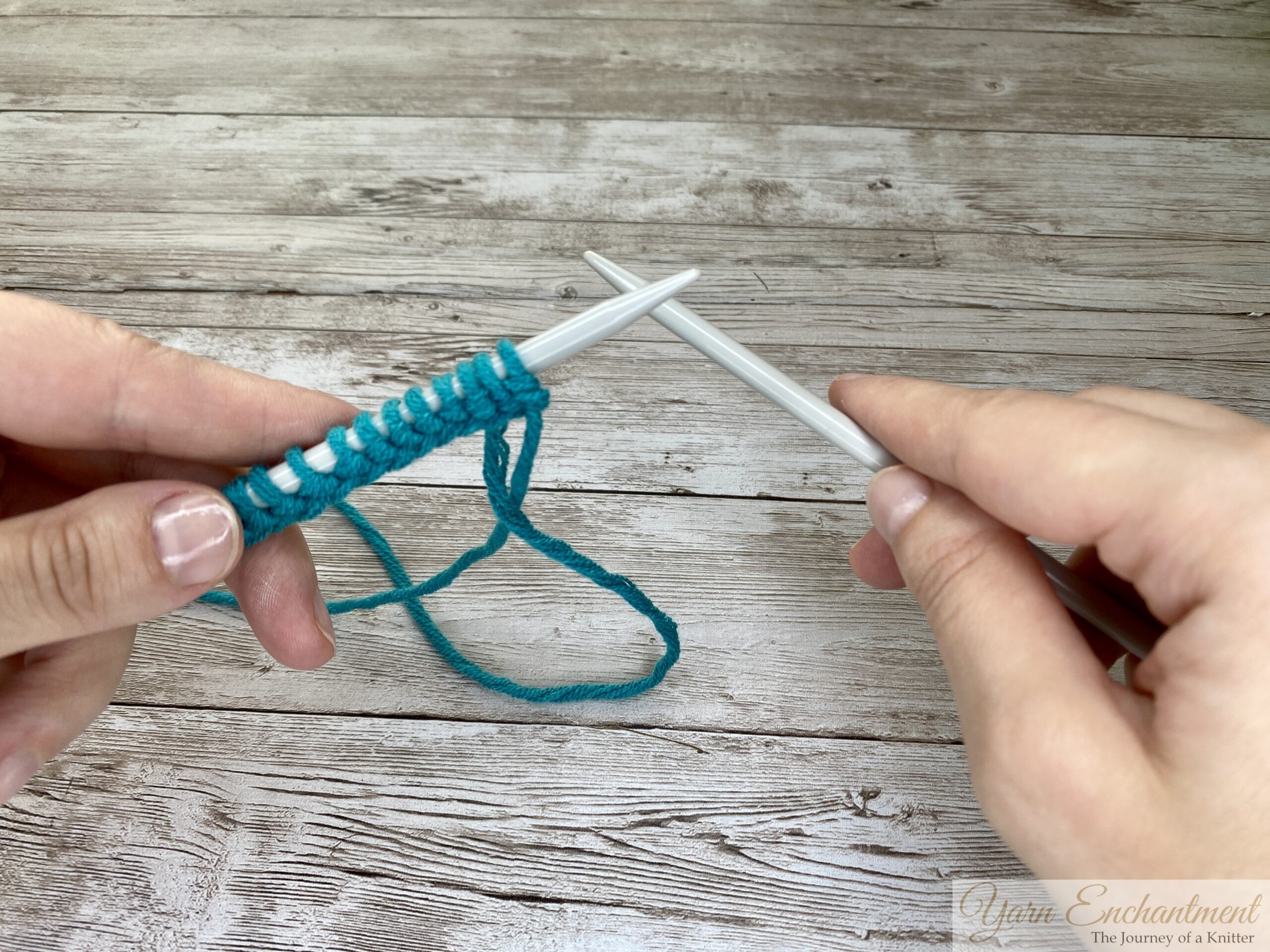 Hands holding knitting needles and a row of turquoise stitches, preparing to begin the first knit or purl row after casting on, with yarn visible on a light wooden surface.