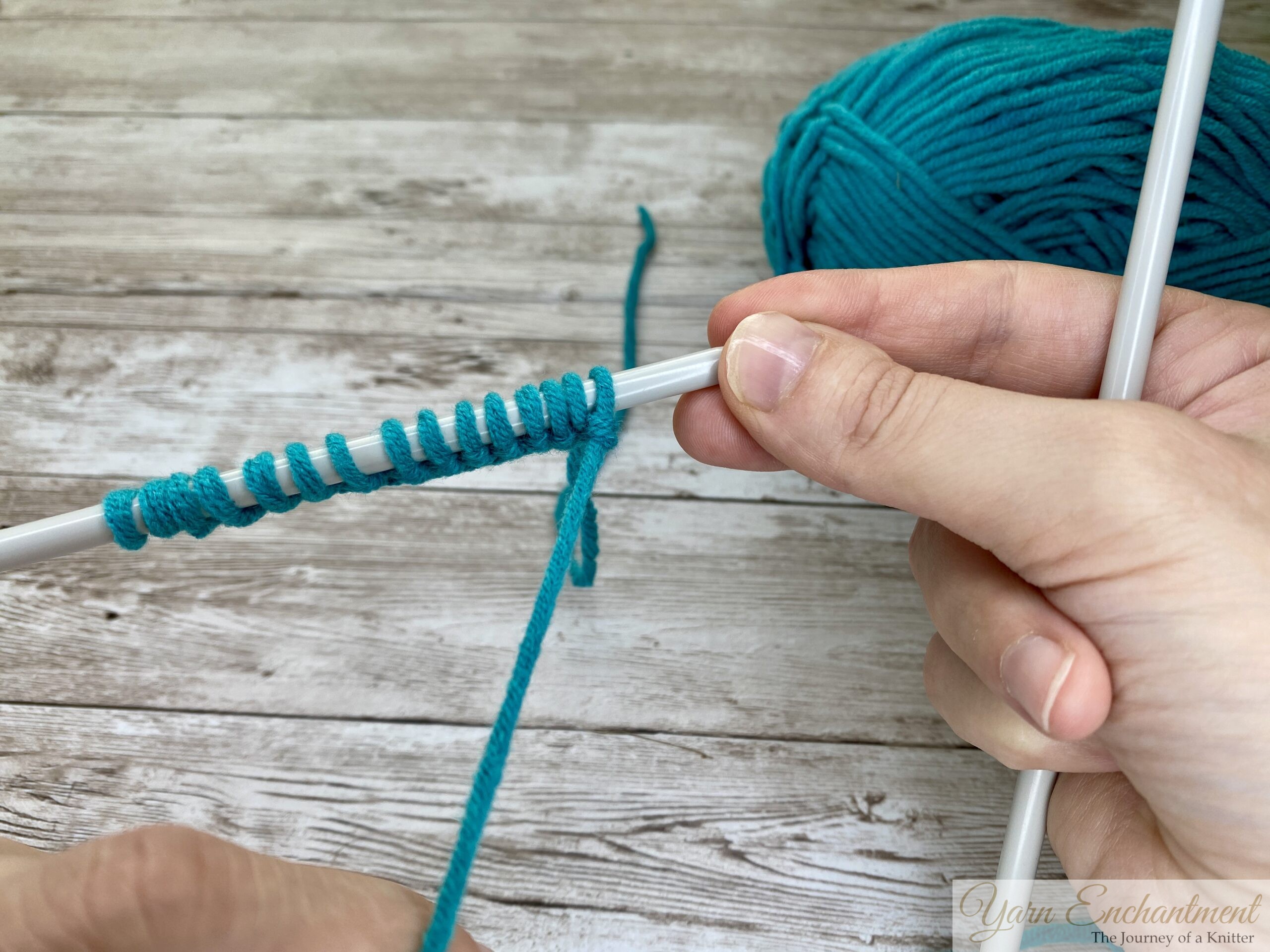 Close-up of hands casting on turquoise yarn onto a knitting needle, showing the start of a knitting project with a ball of yarn in the background.
