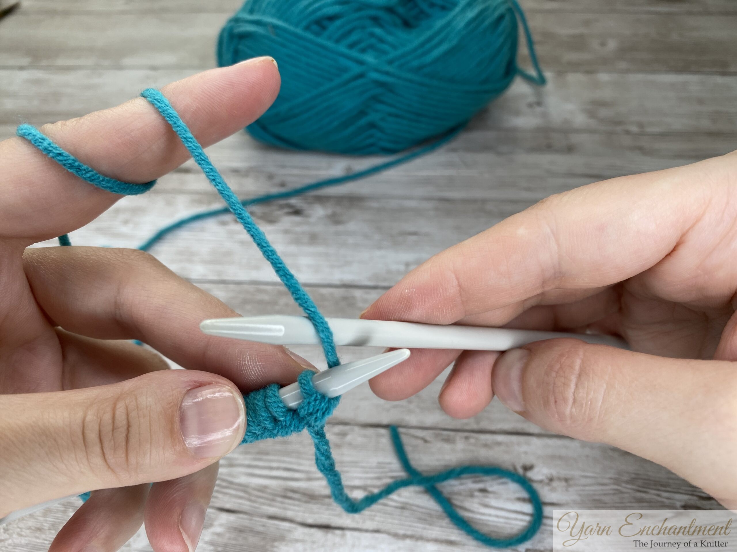 Close-up of hands bringing turquoise yarn to the front of the work with a light gray knitting needle, preparing to position the yarn for the next stitch