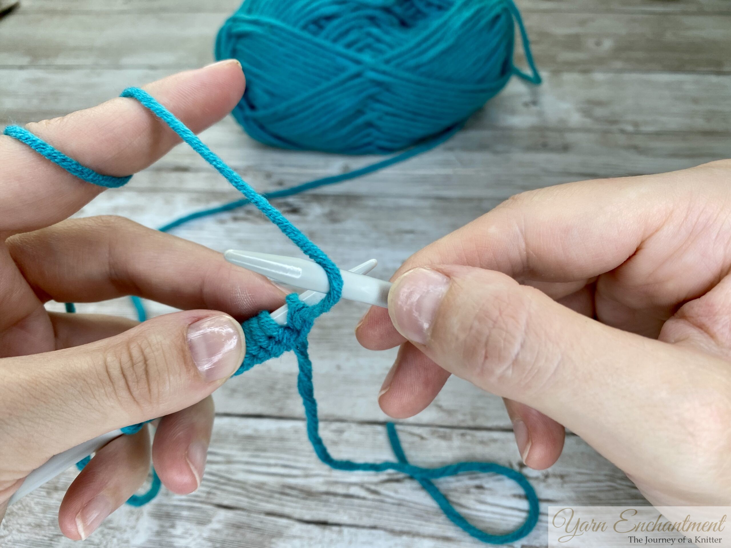 Close-up of hands bringing turquoise yarn to the front of the work with a light gray knitting needle, preparing to position the yarn for the next stitch