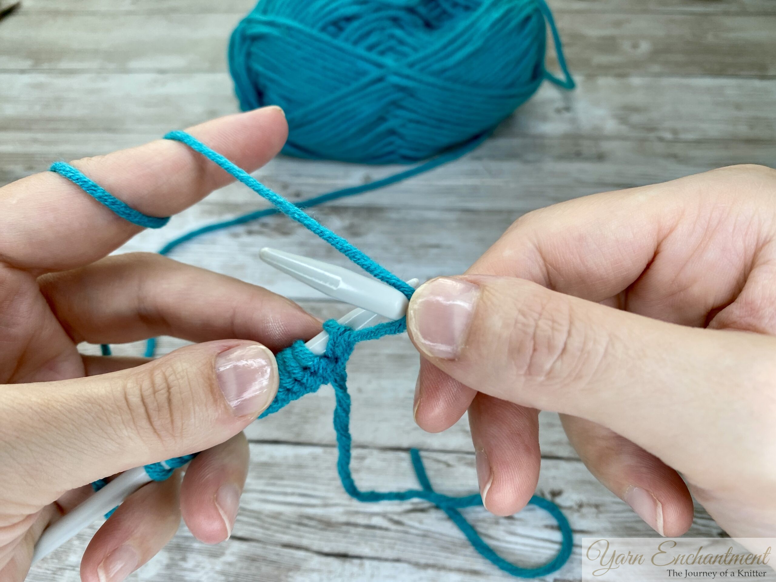 Close-up of hands securing turquoise yarn on a light gray knitting needle using a thumb for stability, ensuring the yarn is ready for knitting