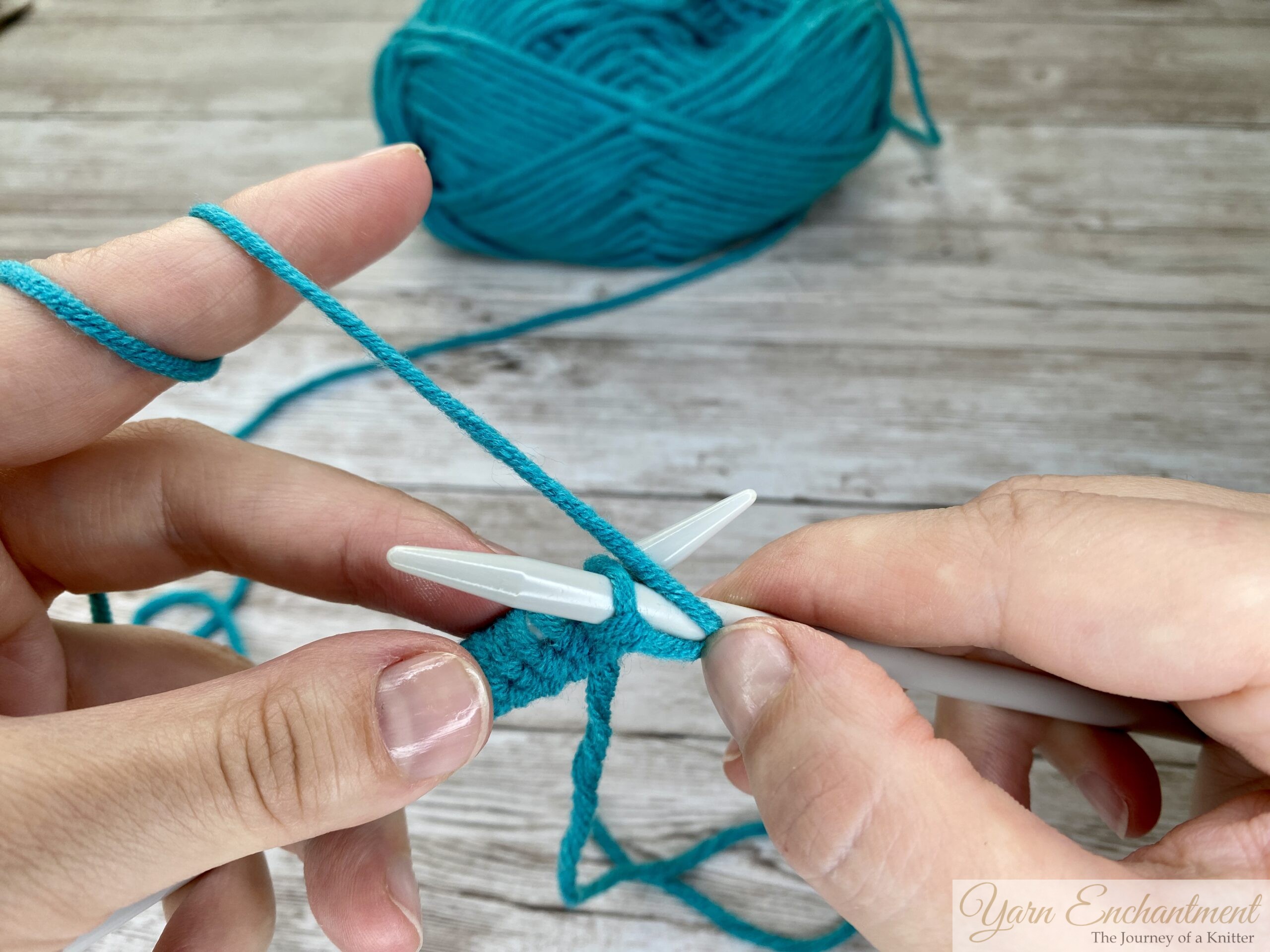 Close-up of hands inserting the right knitting needle from back to front (purlwise) into the first stitch on the left needle, with turquoise yarn and light gray needles on a wooden surface.