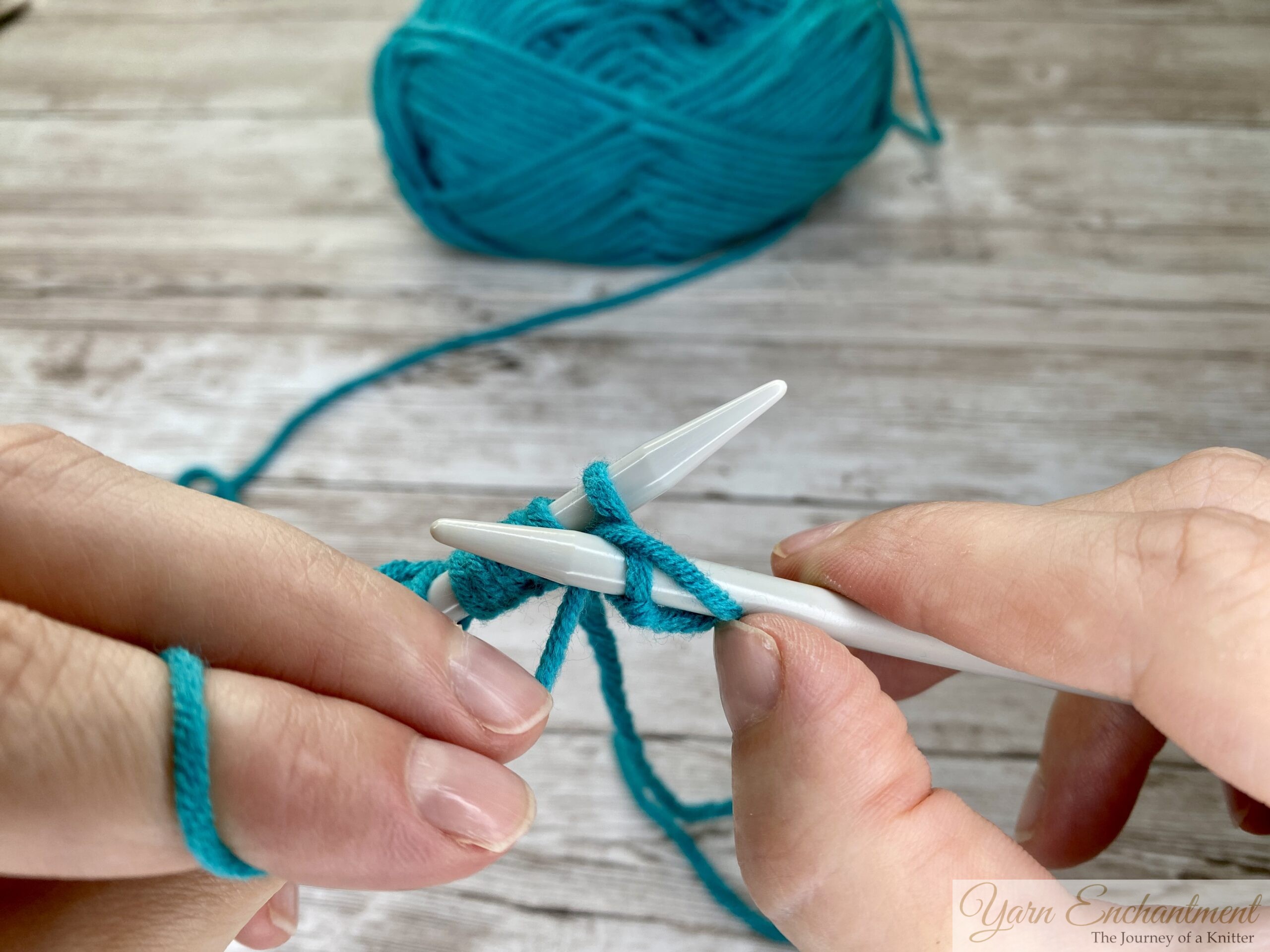 Close-up of hands wrapping turquoise yarn counterclockwise around the tip of the right knitting needle after inserting it into the first stitch, with light gray needles on a wooden surface.