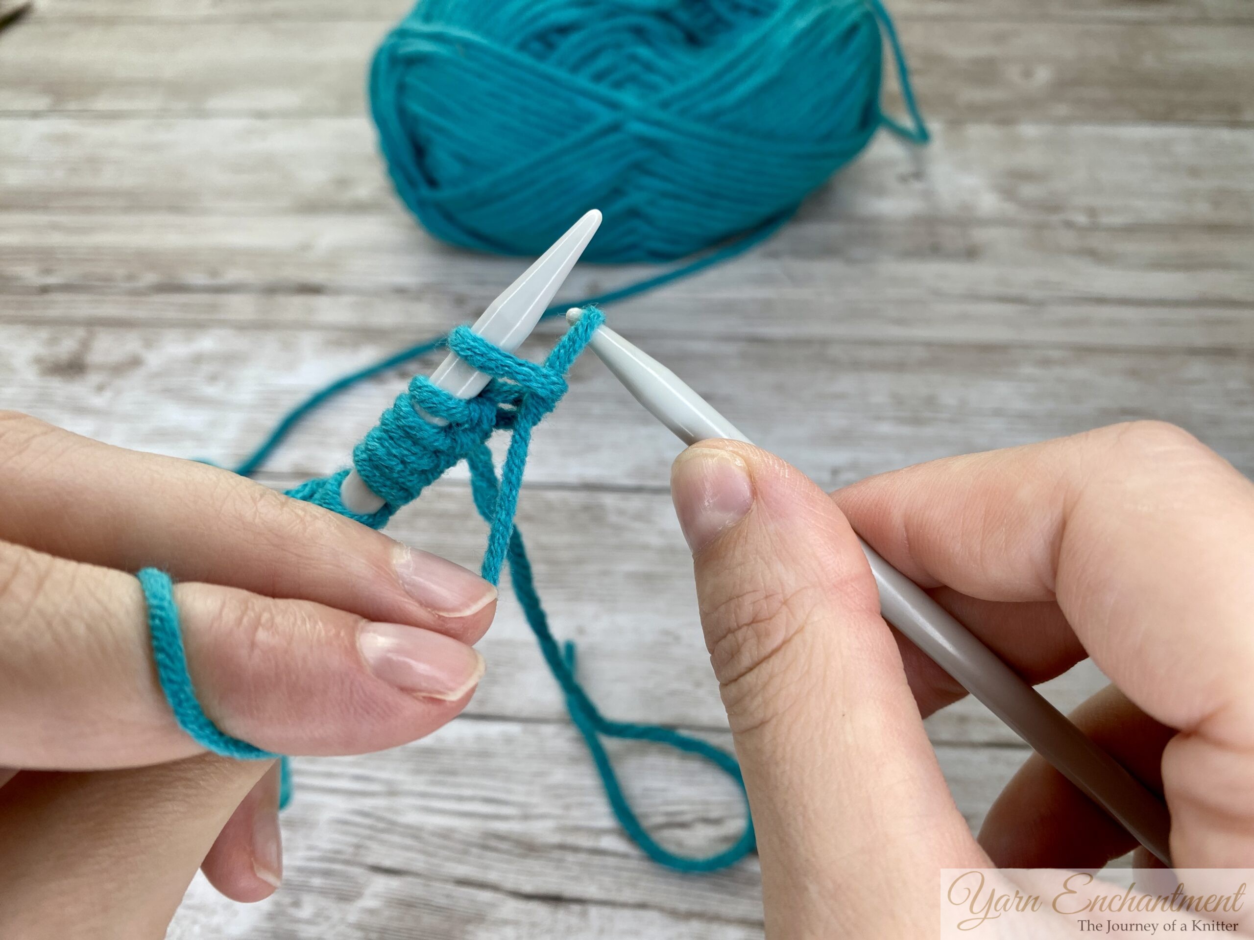 Close-up of hands pulling turquoise yarn through the stitch on a light gray knitting needle to create a new stitch, with the wooden surface in the background