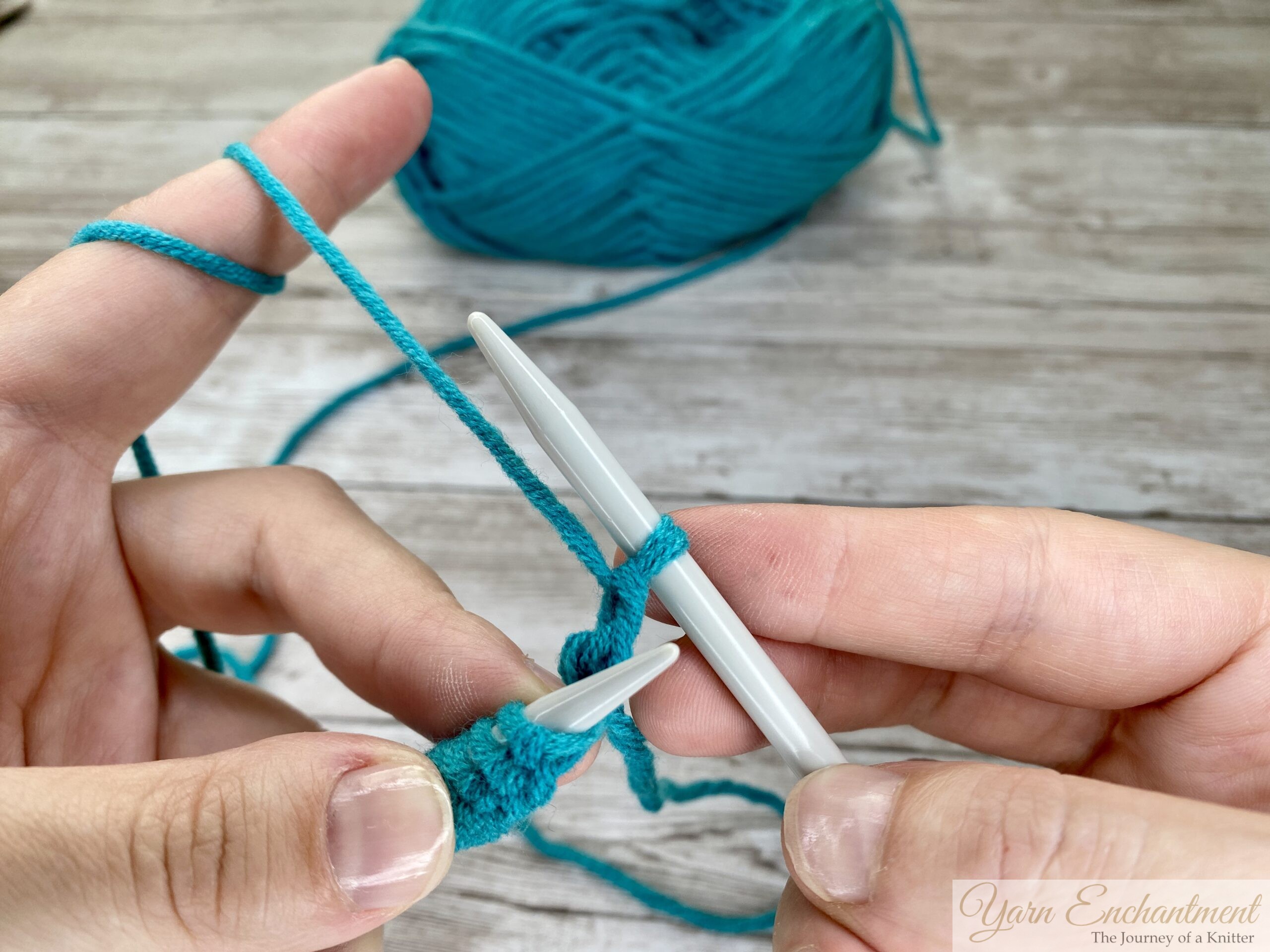 Close-up of hands showing a newly created purl stitch on the right knitting needle, with turquoise yarn and light gray needles on a wooden surface