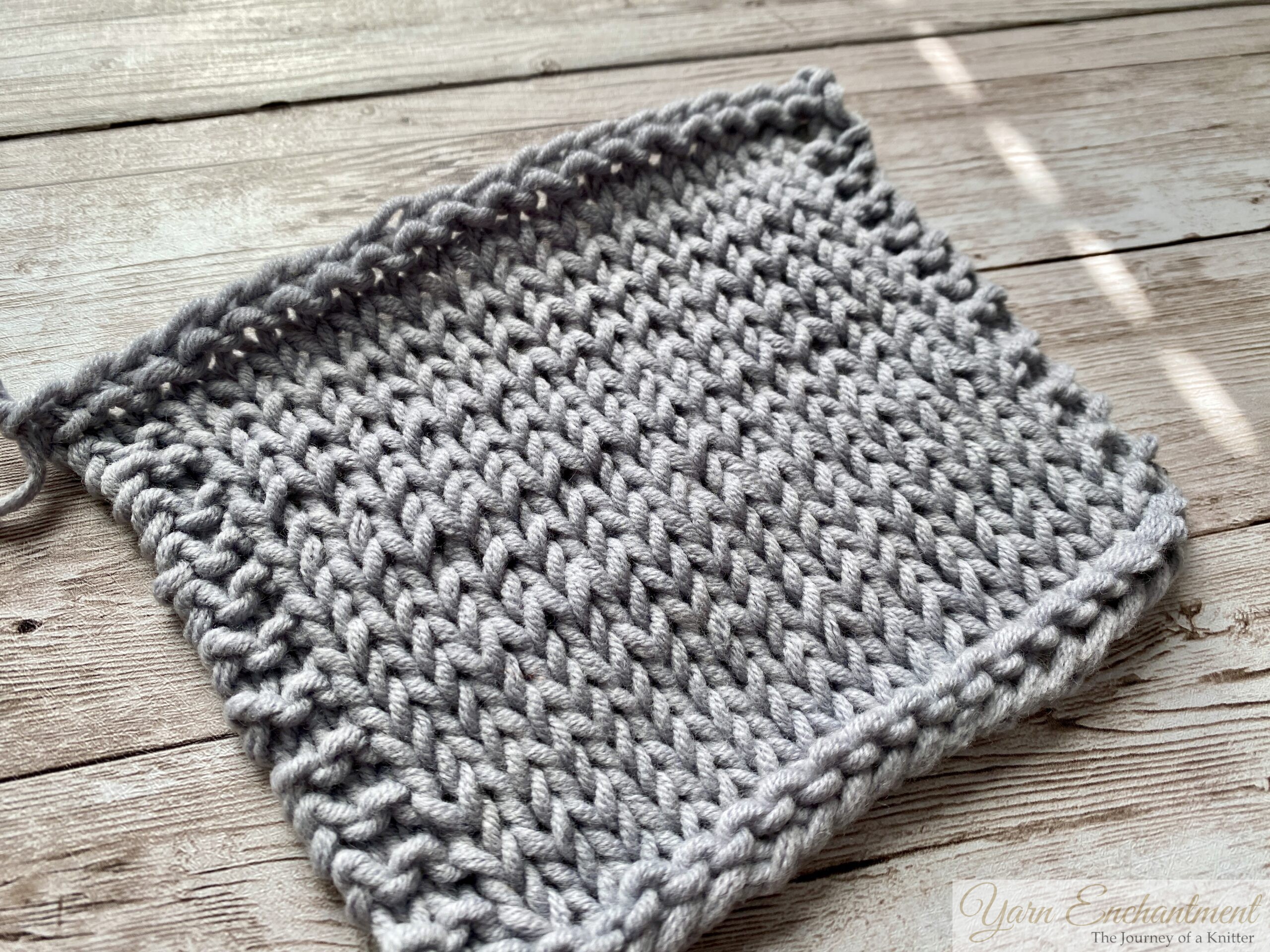 A square swatch of light gray knitted fabric in stockinette stitch, showing uneven horizontal stripes caused by rowing out. Some rows of stitches are noticeably looser, creating subtle ridges across the smooth side of the fabric. The swatch is displayed on a wooden surface.