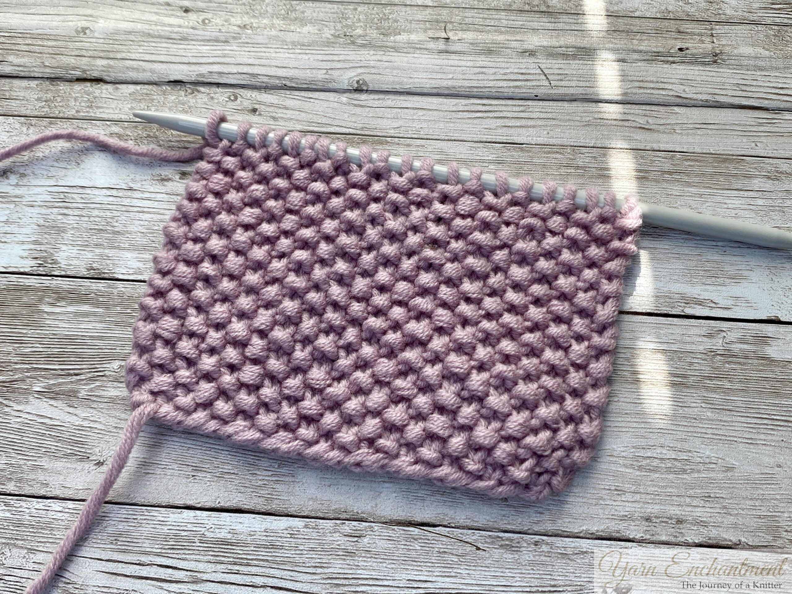 A small, rectangular knitted swatch in soft lavender yarn showing the moss stitch pattern, with visible textured bumps. The swatch is on a single straight knitting needle, resting on a light wooden surface. The working yarn extends from the left side, and each stitch is clearly defined.