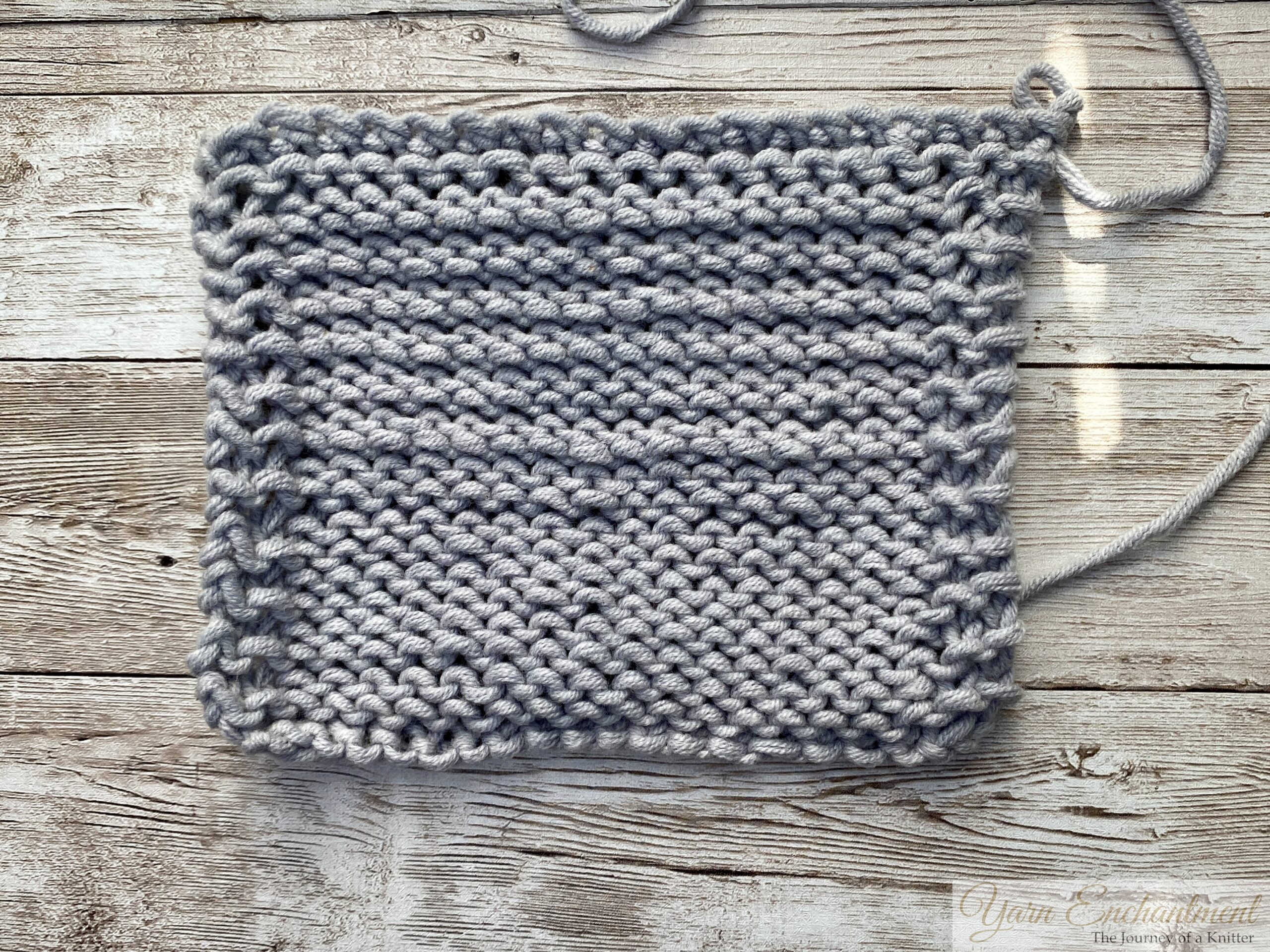 A square swatch of light gray knitted fabric viewed from the purl side, displaying pronounced horizontal ridges from uneven tension (rowing out). The bumps of the purl stitches look irregular across the swatch. The knitting is photographed flat on a wooden background.