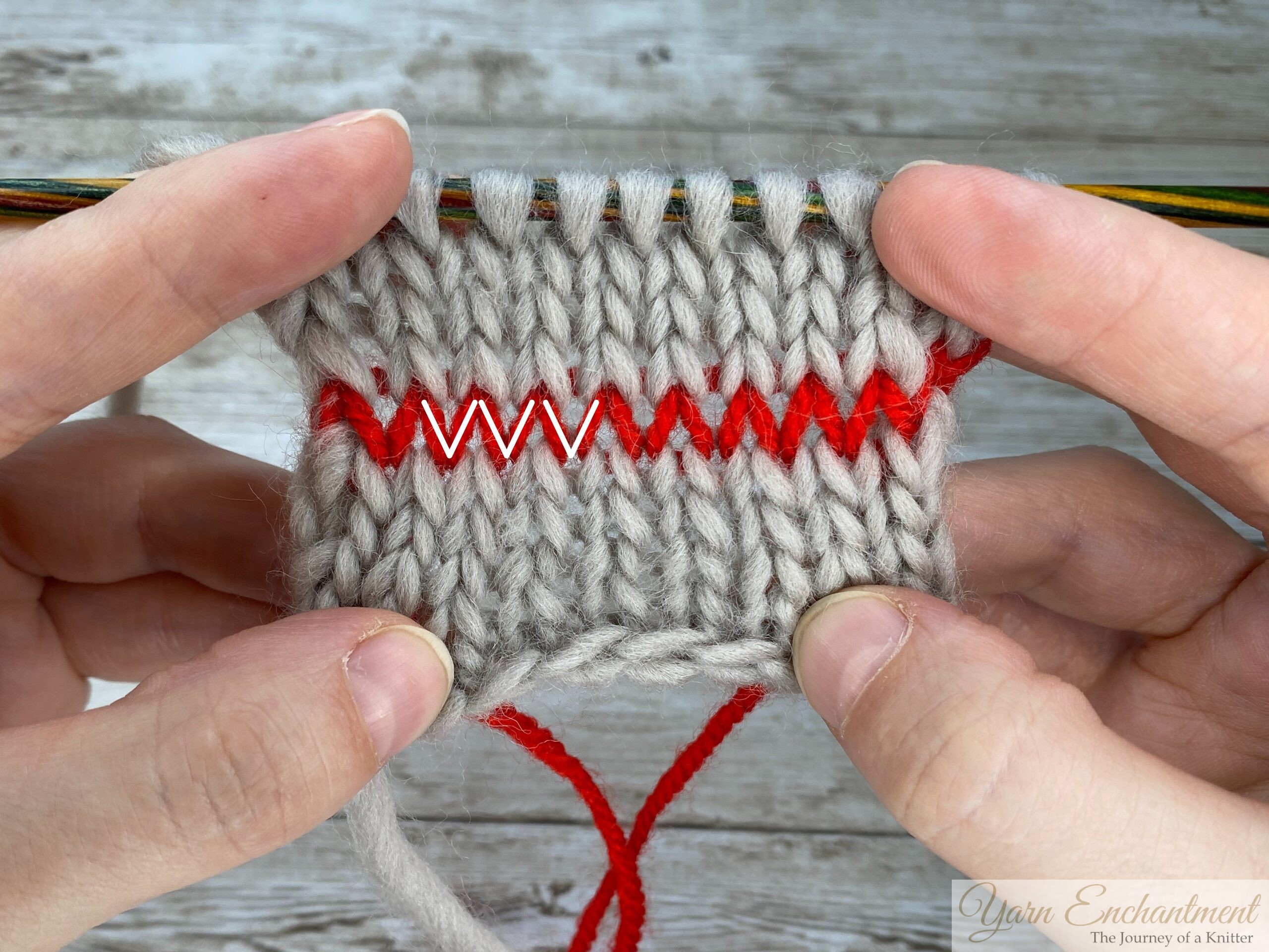 Hands holding a piece of gray knitting on needles, with a row of red contrasting yarn woven horizontally through the stitches, clearly highlighting the “V” shape of knit stitches for instructional purposes.