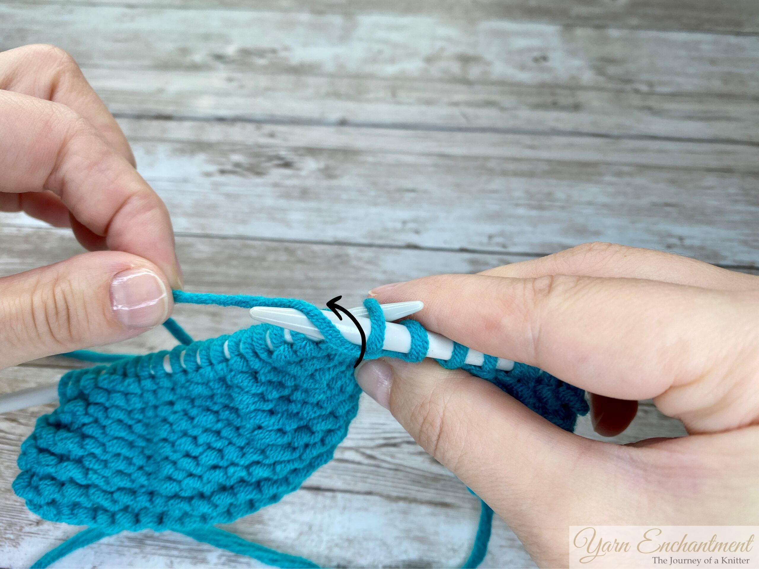 Close-up of hands wrapping turquoise yarn counterclockwise around the tip of a light gray knitting needle, with turquoise yarn and light gray knitting needles on a wooden surface background.