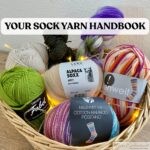 A woven basket filled with colorful skeins and balls of sock yarn—including green, purple, cream, and variegated options—arranged on a wooden surface, with the overlaid title “YOUR SOCK YARN HANDBOOK” in bold black text on a white banner above the yarn.