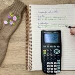 Knitted sock with embroidered flowers next to a notebook showing sock cast-on stitch formulas in inches and centimeters, a TI-82 calculator, and a hand writing with a pen.