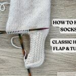 In-progress cream-colored hand-knit sock on double-pointed needles, showing a finished heel flap and turned heel on a wooden surface, with the text 'How to Knit Socks: Classic Heel Flap & Turn' in bold black letters on a white background.
