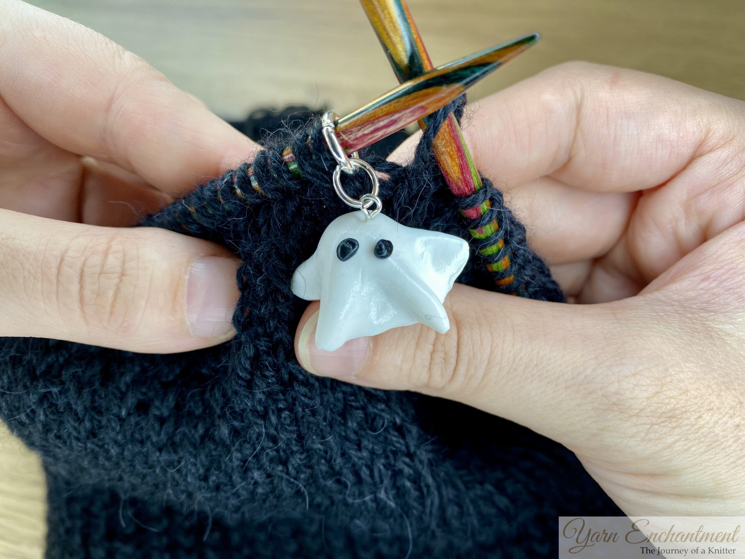 Hands knitting with dark yarn on circular needles, using a cute ghost-shaped stitch marker to track progress and add fun to the project.