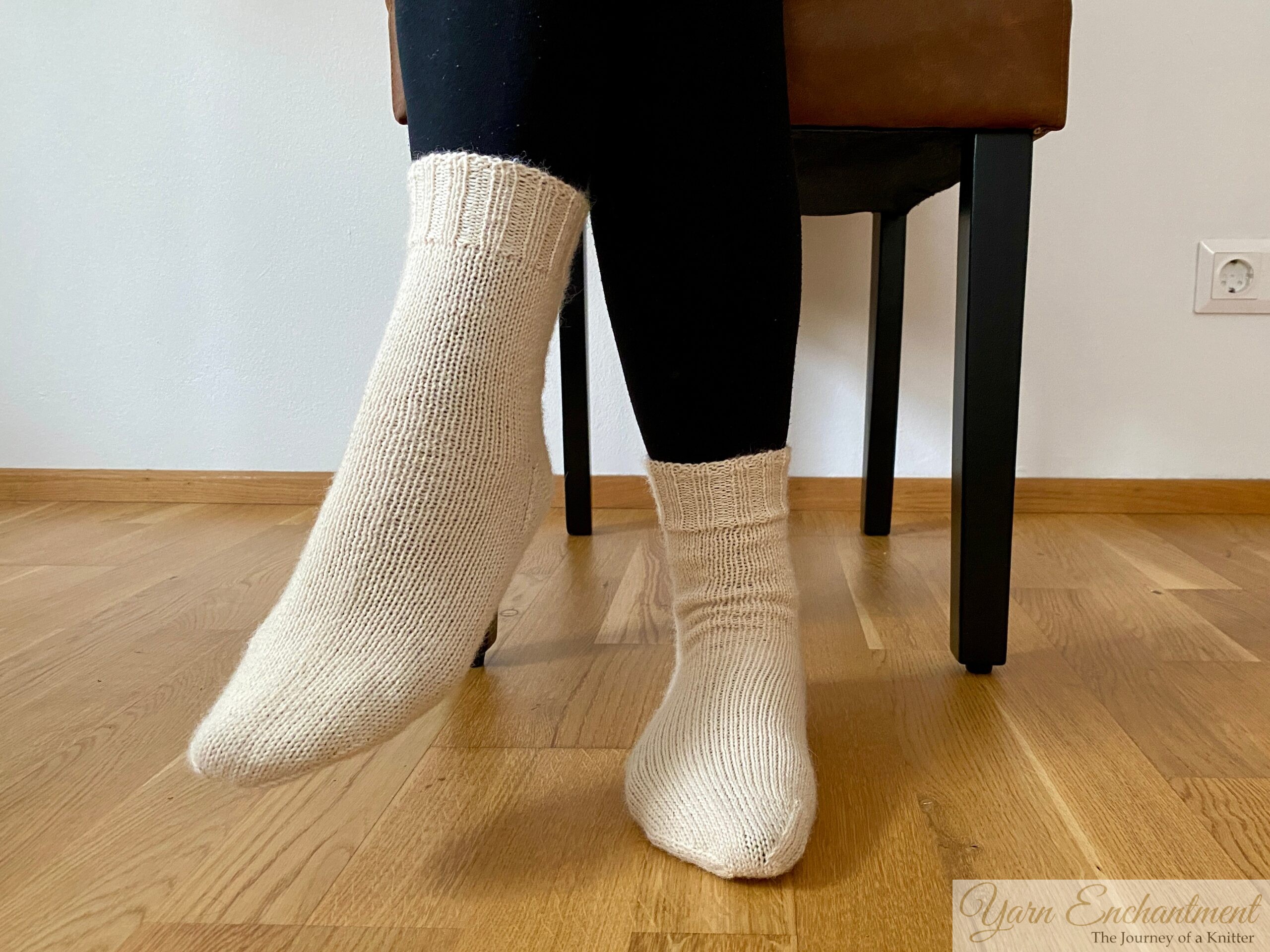 Hand-knitted cream-colored socks on feet, shown from a side angle while standing on a wooden floor. The socks are paired with black leggings, and the photo focuses on showcasing the finished texture and fit of the socks.