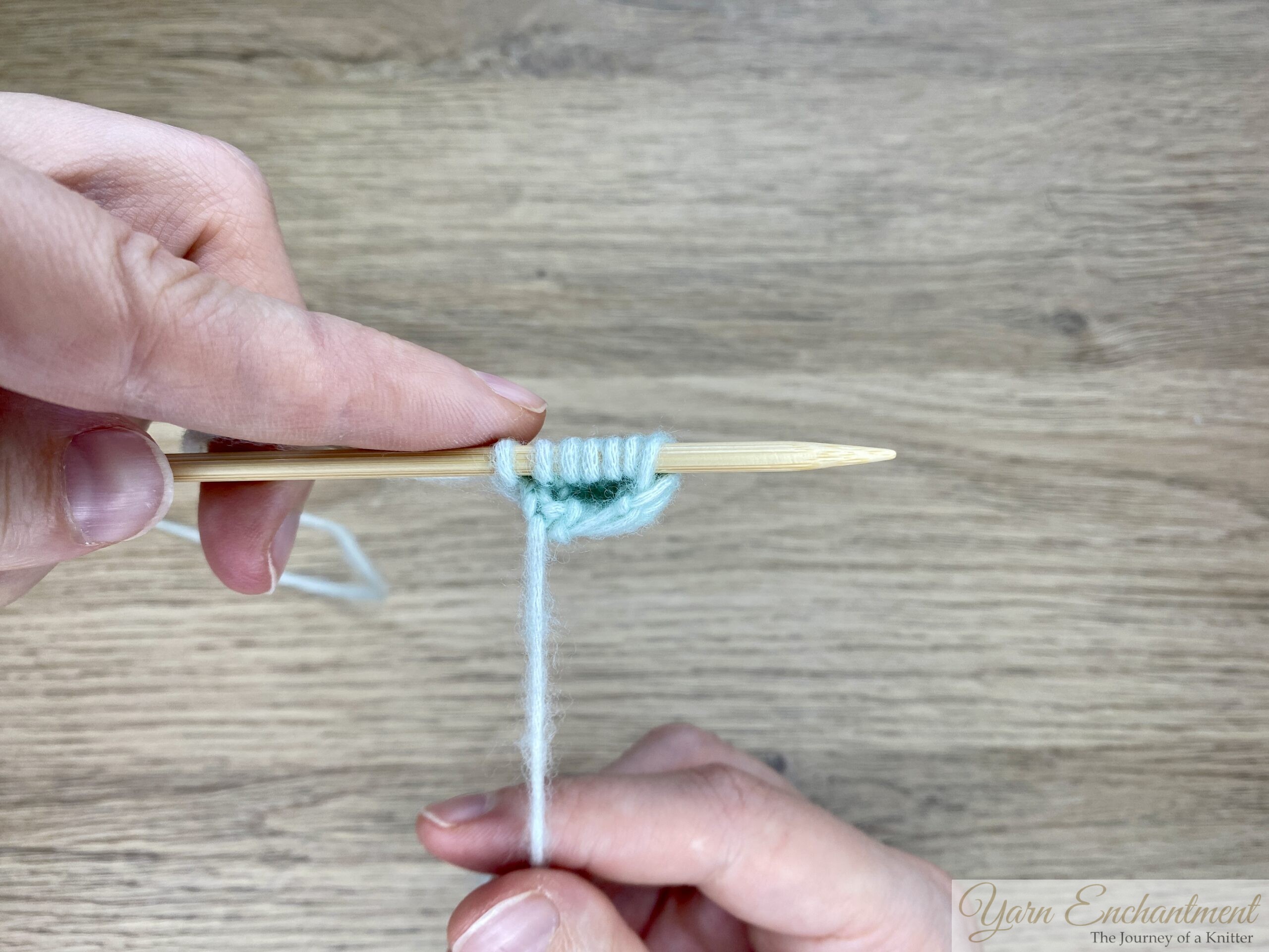 Fingers lightly pressing on a bamboo knitting needle with several stitches of mint green yarn; the I-cord is shown from the side, highlighting the tube-like shape.